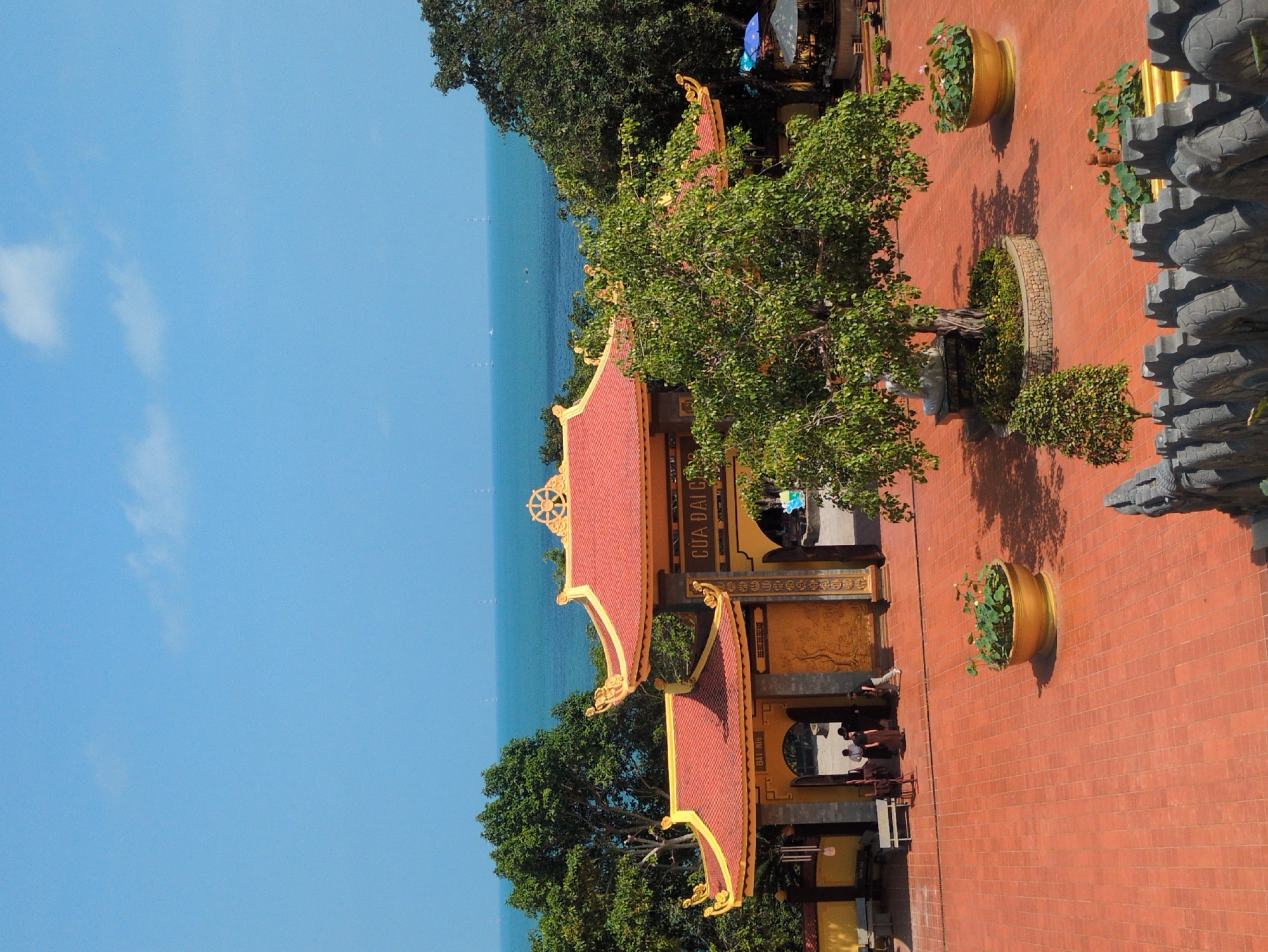 The image shows a view of the Cua Dai Cac Bat Nha temple in Phú Quốc, Vietnam, taken from an elevated position. The temple structure features a red-tiled roof with golden trim. The structure bears the inscription "CUA DAI CAC BAT NHA". Trees surround the temple complex, and the ocean is visible in the background. The foreground displays a brick-paved area with decorative stone elements and potted plants. People are visible, reflecting in a glass panel.