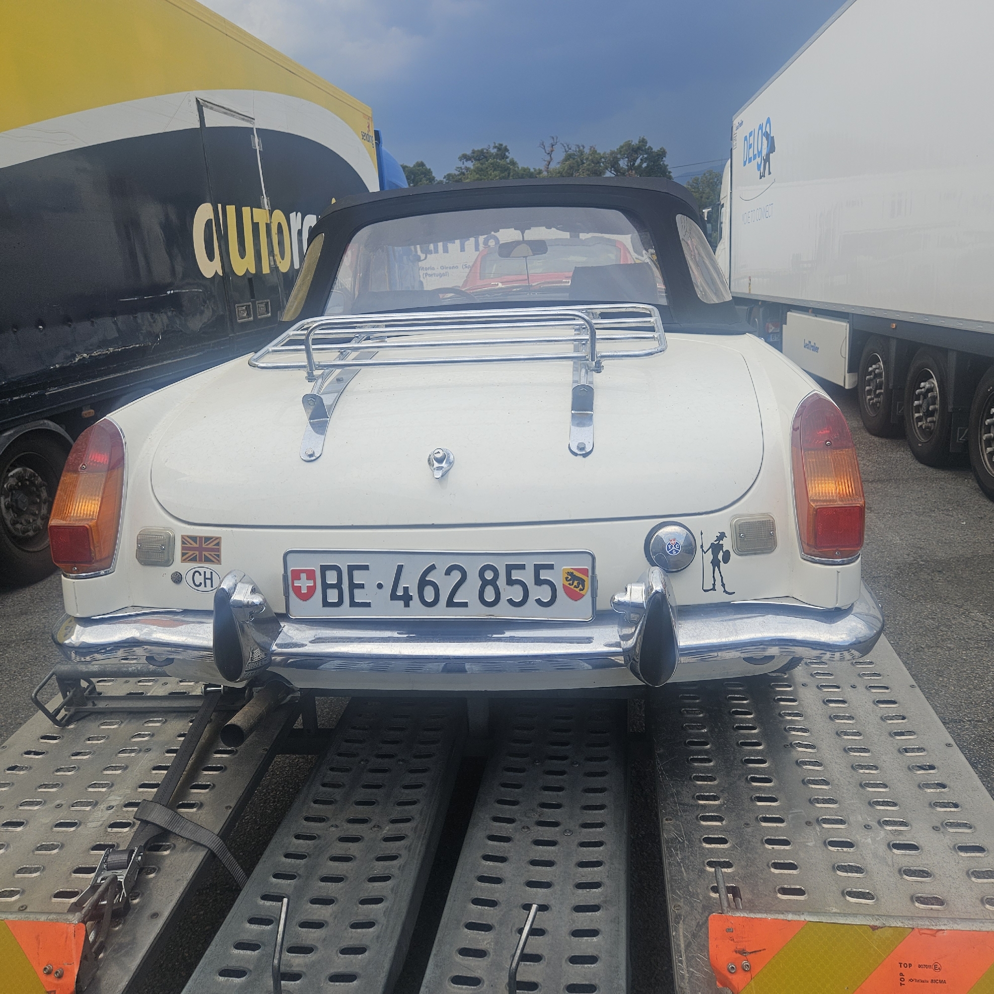 The image shows the rear of a white convertible car being transported on a trailer. The car has a Swiss license plate that reads "BE 462855". There are stickers on the car, including a Swiss flag, a Union Jack, and a figure of a man with a spear. The car is parked next to two large trucks. One truck is yellow and black with the word "autoropa" printed on it. The other truck is white with the logo "DELGOS" and the words "NO TO CONNECT" visible. The scene appears to be a parking area, possibly a rest stop or logistics hub, given the presence of multiple transport vehicles. The sky is cloudy, suggesting overcast weather. The location is identified as La Jonquera, Spain.