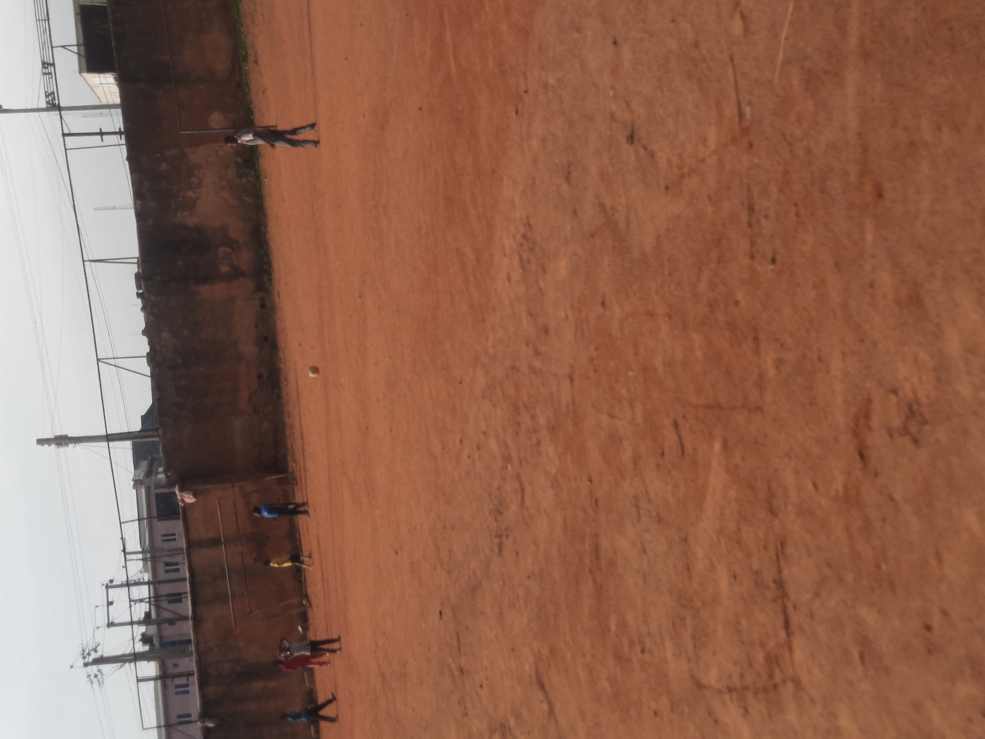 The scene depicts an open dirt field in Jos, Nigeria. Several people are present, spaced along the edge of the field adjacent to a wall. Overhead power lines and utility poles are visible, as are structures including buildings. One person appears to be running or walking across the field. A small, light-colored sphere lies on the field.