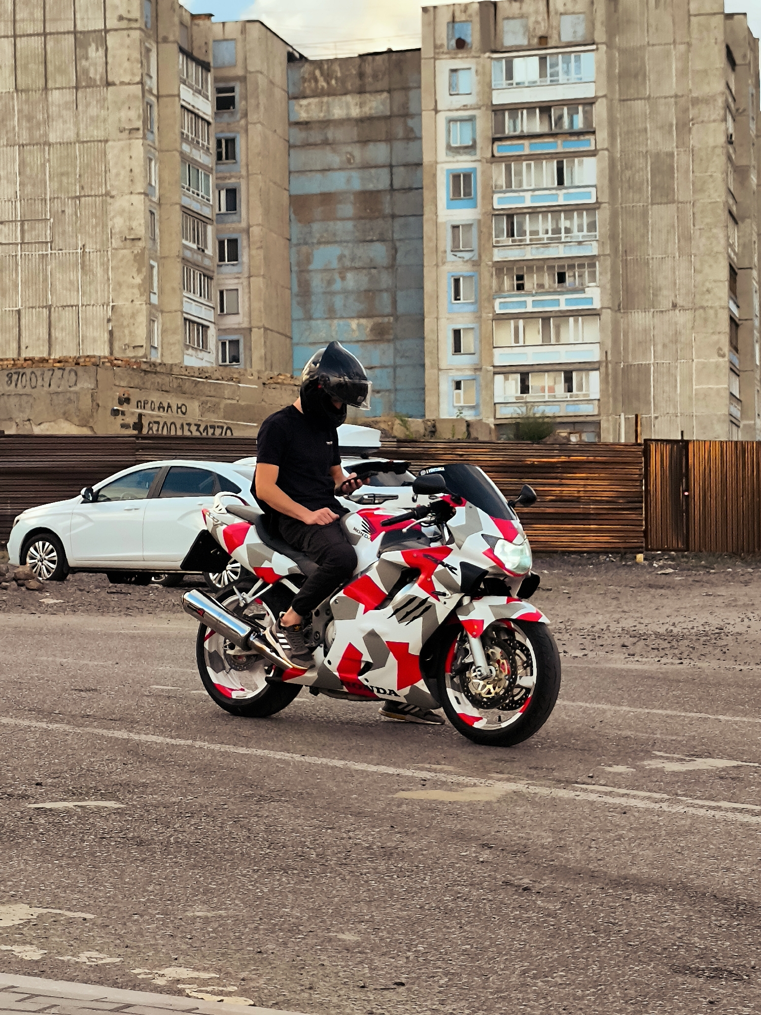 In Qarağandı city administration, Kazakhstan, a person in a black helmet and black clothing is seated on a white, red, and gray camouflage motorcycle. The motorcycle is parked on a gray asphalt surface. In the background, there is a white car and a multi-story building with weathered concrete walls and blue-accented windows. A wooden fence is visible behind the car. The scene appears to be an outdoor urban environment during daylight.
