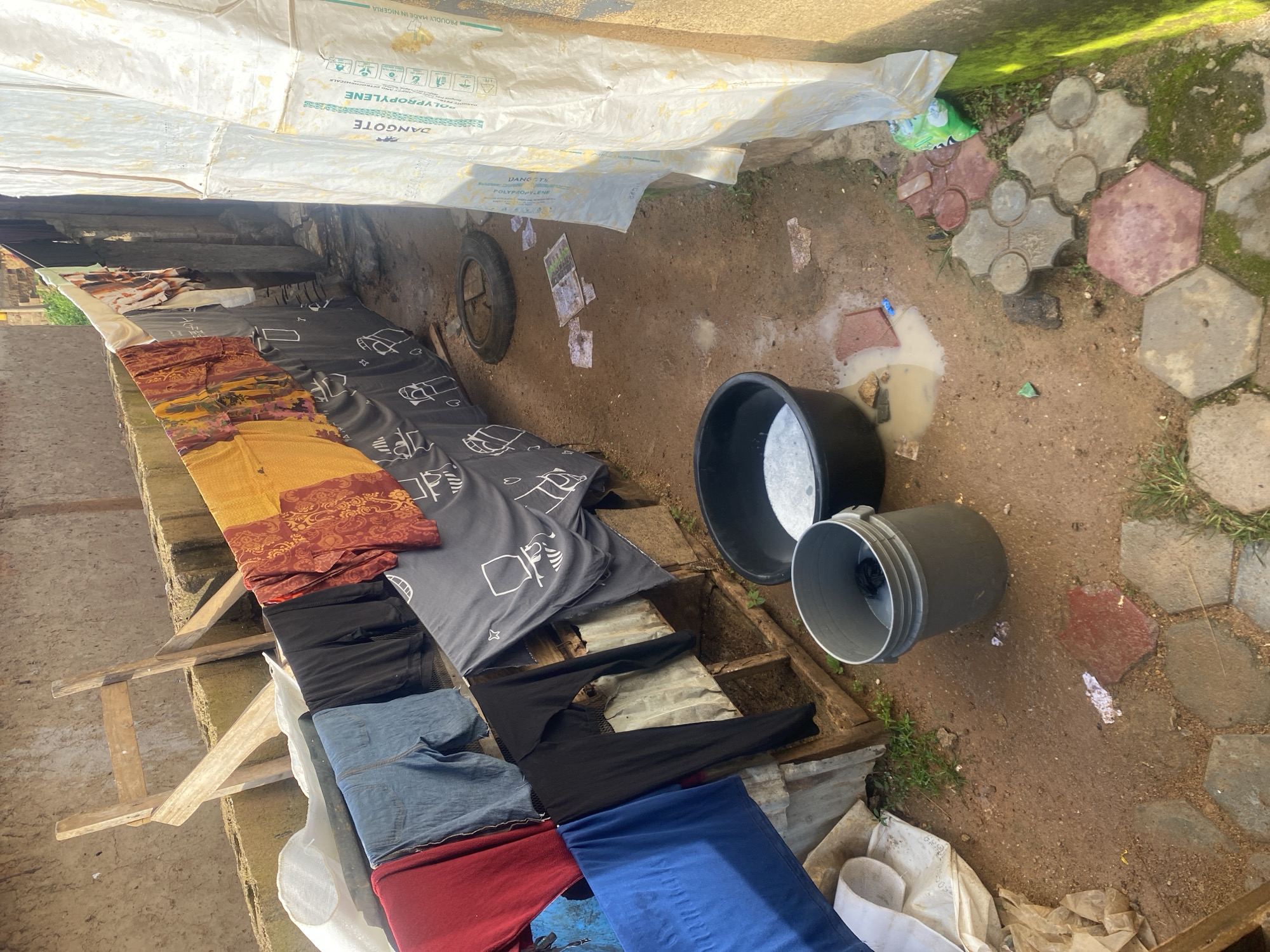 The image depicts an outdoor area, likely a backyard or alleyway, in Kabong, Nigeria. Clothes are hung on a clothesline, including a red garment, blue denim shorts, black leggings, and patterned fabric in yellow and red. Some of the clothes have simple graphic designs on them. Below the clothesline, there are some wooden structures and what appears to be corrugated metal.
On the ground, there are two buckets. One is a large black basin filled with a white, soapy liquid, suggesting laundry is in progress. Beside it is a gray plastic bucket containing some dark items and a small amount of liquid. The ground is dirt with scattered debris, including some papers and a worn tire. Puddles of water are visible, indicating recent rain or washing activities.
To the right of the scene, a large white tarp or plastic sheeting is propped up against a wall. The tarp has text and symbols printed on it, including "DANGOTE POLYPROPYLENE." The wall appears to be made of concrete blocks and is partially covered in green moss. The ground in the foreground features interlocking hexagonal and clover-shaped pavers. The overall impression is of a utilitarian, possibly low-income, residential setting where everyday chores are being carried out.