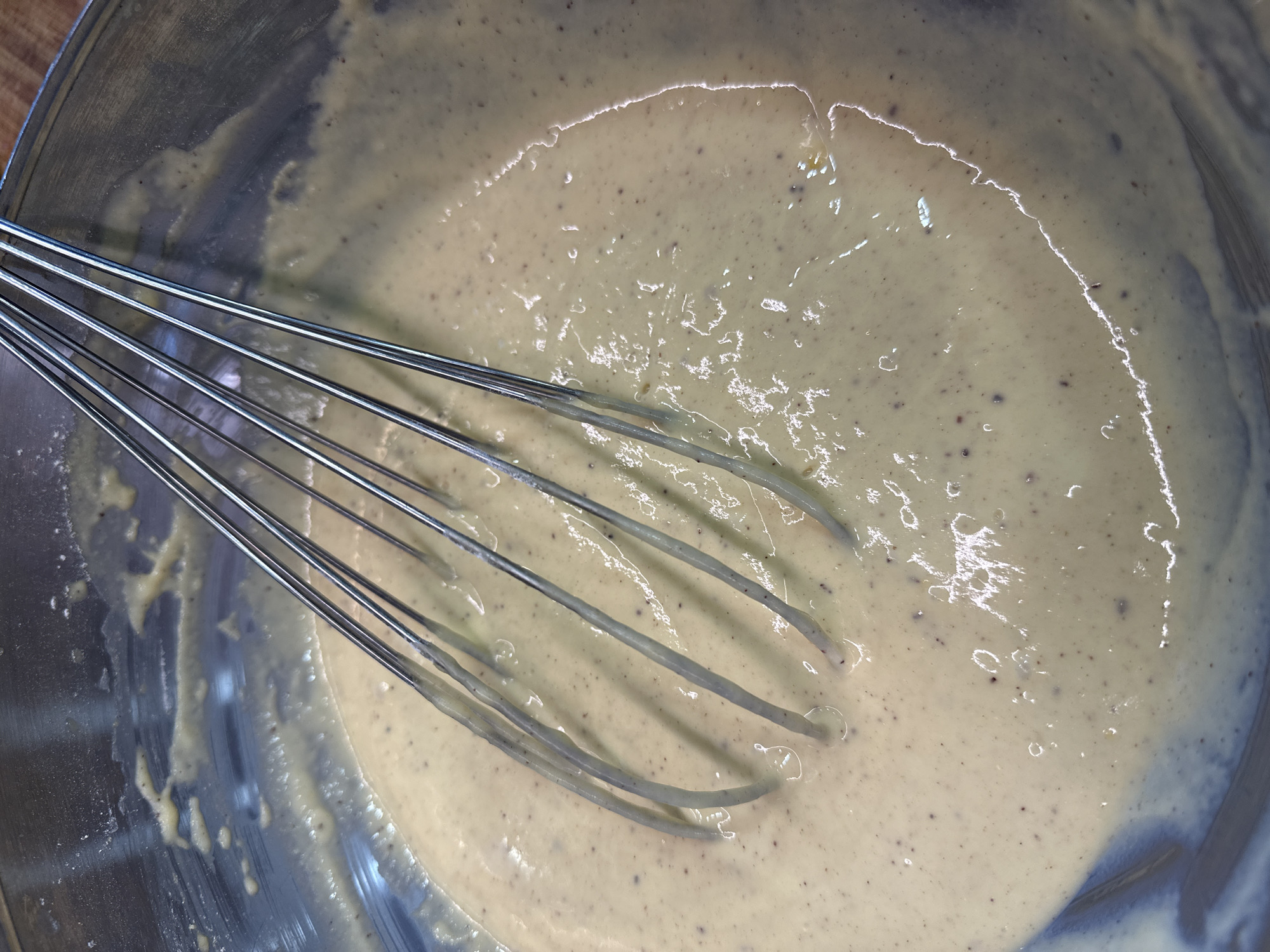 A close-up overhead shot shows a whisk submerged in a pale yellow batter within a silver metal bowl. The batter is thick and appears to contain small dark specks. The whisk, made of thin metal wires, is partially coated with the batter. The bowl's surface shows reflections and some smudges. The background is blurred, suggesting a kitchen environment. This image is not related to Nevinnomyssk, Russia.