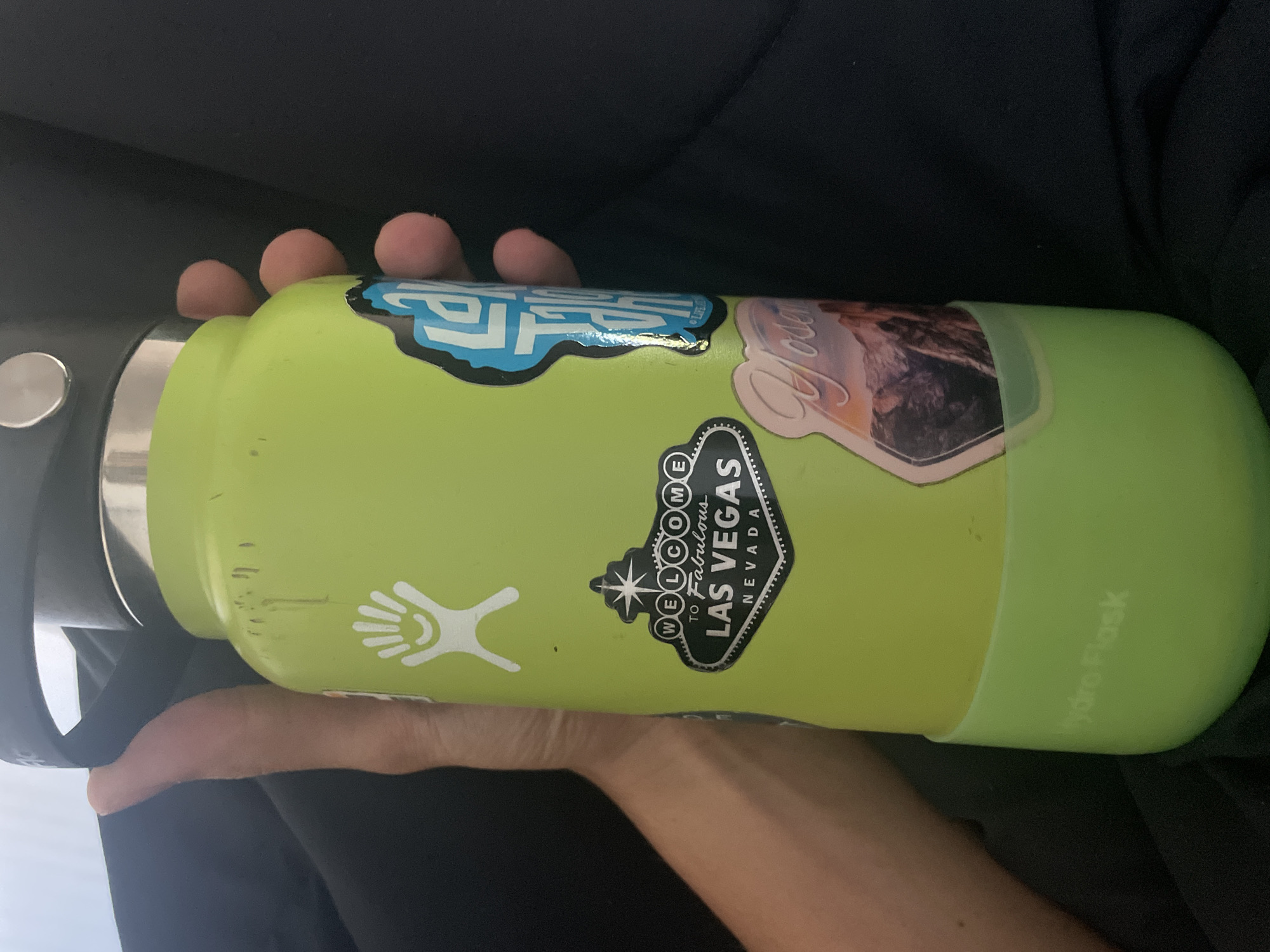 A person is holding a green Hydro Flask water bottle. The bottle has several stickers on it, including a "Welcome to Fabulous Las Vegas, Nevada" sign sticker and a white hand logo sticker. The person's hand and arm are visible. The background is dark and out of focus.