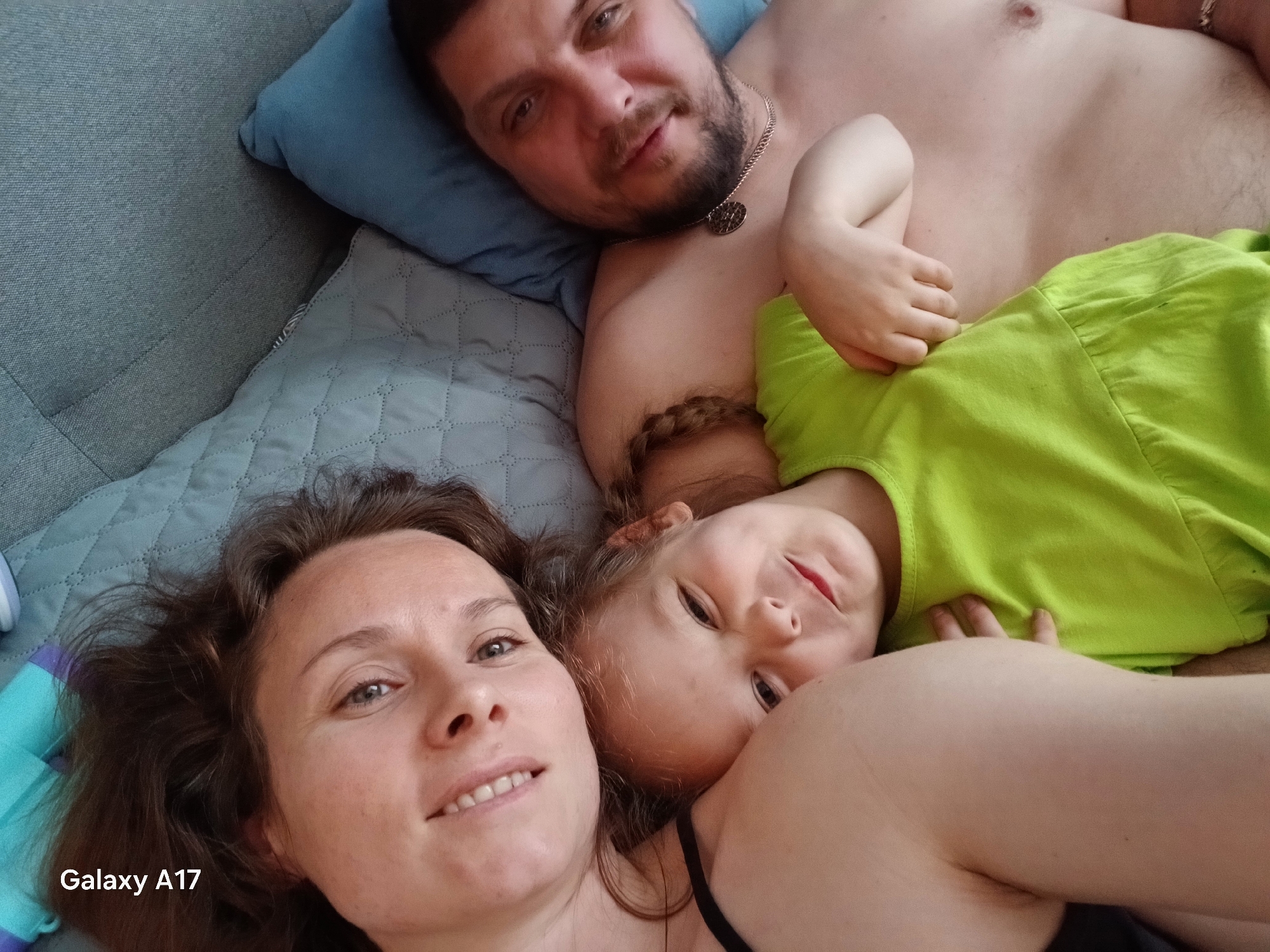 A selfie of a family of three taken indoors. A woman is in the foreground with brown hair and a black tank top. To her right is a young girl with blonde braids, wearing a green dress. A man with a beard and chest tattoo is behind them. They are lying on a grey quilted blanket and blue pillow, appearing relaxed. The camera watermark "Galaxy A17" is visible in the lower left corner. The location is not identifiable as Yekaterinburg, Russia from the image.