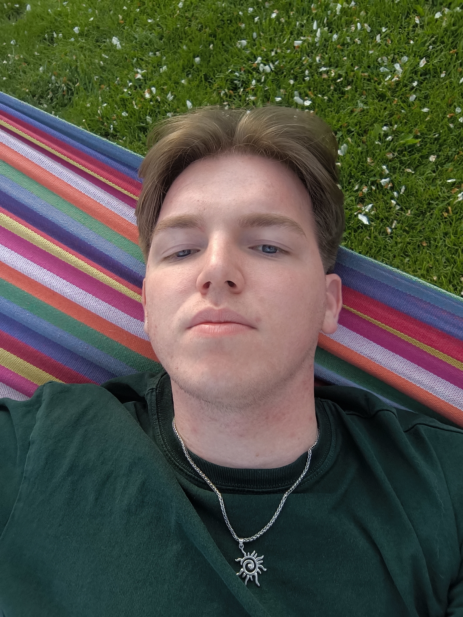 A young man is lying in a colorful hammock outdoors. The hammock has stripes of various colors including pink, purple, orange, and blue. The background shows green grass with white petals scattered on it. The man has fair skin, short light brown hair, and blue eyes. He is wearing a dark green t-shirt and a silver necklace with a sun pendant. The image is taken from a close-up, slightly above angle. The location is likely a grassy area with trees, based on the scattered petals, but no buildings are visible. The specific city or location of Busmantsi, Bulgaria, cannot be verified from the image alone.