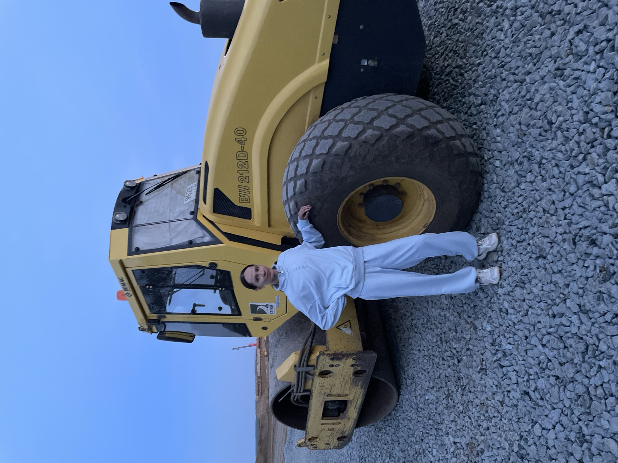 A person dressed in a light-colored tracksuit is standing next to a yellow Bomag BW 212D-40 soil compactor, leaning against one of its large tires. The compactor is parked on a gravel-covered surface. The background includes a blue sky and hints of a construction site. The location is assumed to be in the city of Kabanikha, Russia.