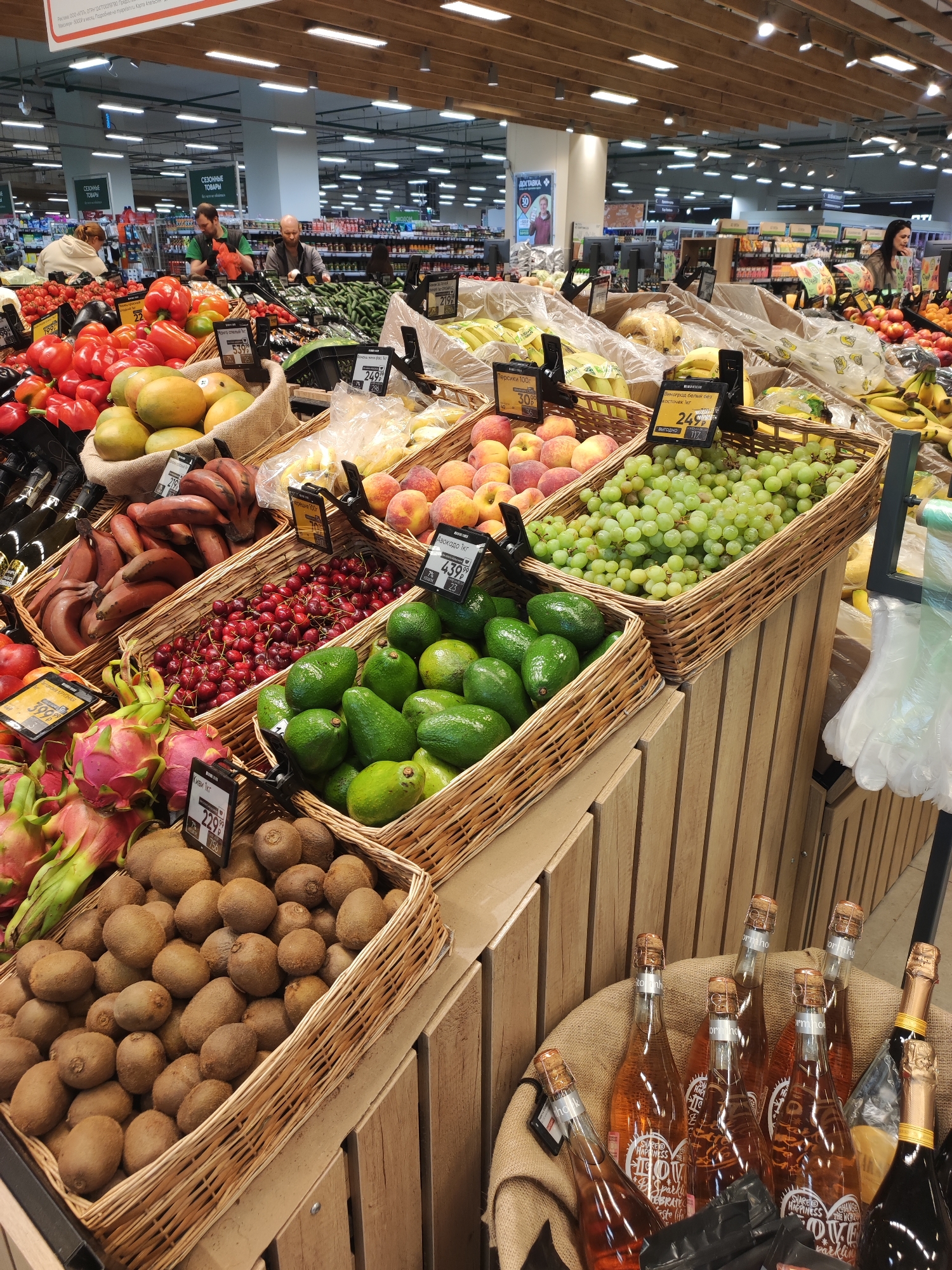 The image captures a colorful and abundant display of fresh produce and beverages inside a supermarket in Sochi, Russia. The scene is dominated by wicker baskets overflowing with a variety of fruits and vegetables, including red bananas, cherries, avocados, grapes, kiwis, dragon fruits, peaches, and mangoes. Price tags are visible, indicating the cost per kilogram in Russian rubles. The produce is arranged on wooden platforms or shelves. In the foreground, there is a burlap sack containing bottles of rosé sparkling wine or similar beverage.
In the background, other grocery items are visible, along with customers shopping in the aisles. Fluorescent lighting illuminates the store. The environment suggests a well-stocked, modern grocery store catering to a diverse clientele.