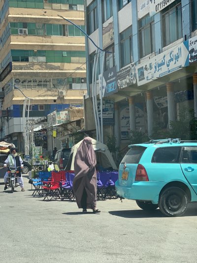 The image shows a street scene in Kandahar, Afghanistan. A woman wearing a full brown burqa stands in the middle of the street. To her right is a light blue car. Behind her are several colorful folding chairs, a white tent, and tables with items on display. To her left, a man rides a motorcycle. Buildings line the street, with signs in Arabic and English.