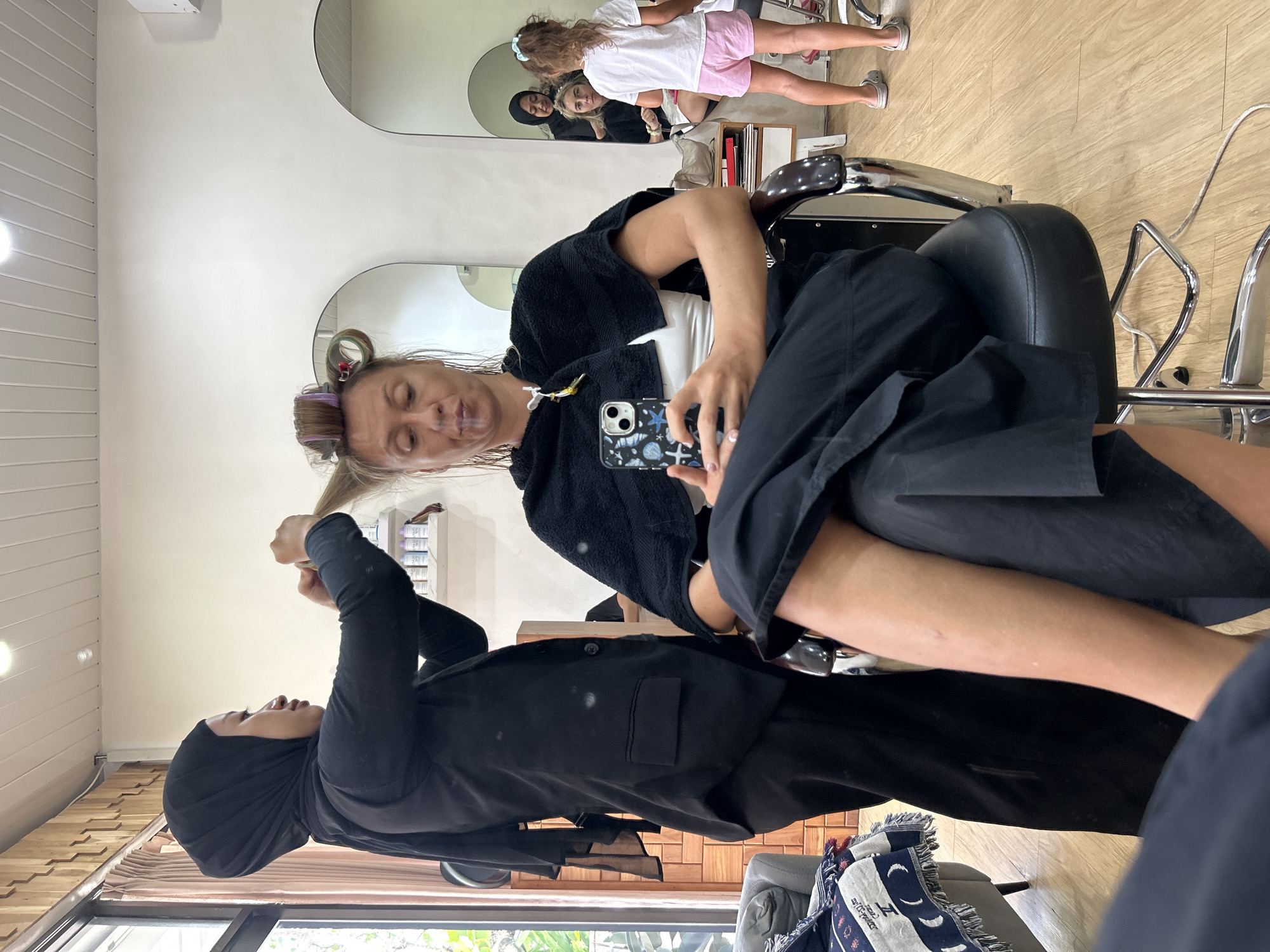 A woman wearing a black hijab and black blazer styles the hair of a client seated in a salon chair in North Kuta, Indonesia. The client, a woman with blonde hair styled in large rollers, wears a white tank top, a black cape, and black pants. She is holding a smartphone in her left hand and appears to be smoking something, possibly a cigarette, with a visible plume of smoke. In the background, a young girl in a white t-shirt and pink shorts stands near a mirror, and another woman is visible in the reflection of the mirror, also in the process of getting her hair done. The salon features wooden flooring, mirrored walls, and white paneling on the ceiling. A sofa with a patterned throw is visible on the left side of the image.