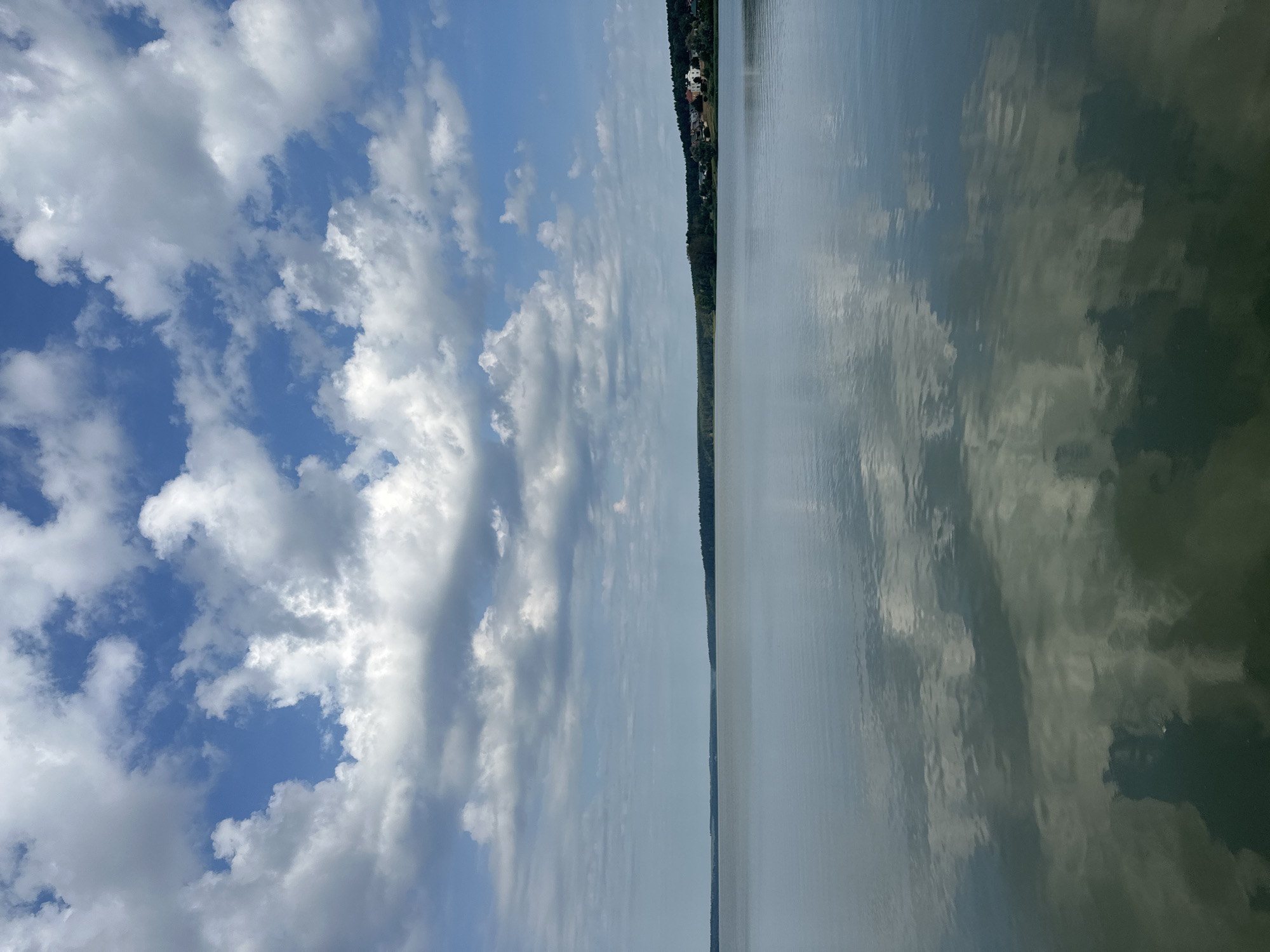 The image depicts a serene landscape on a body of water, likely a lake or river, near the city of Votkinsk, Russia. The sky is filled with a dramatic display of white, cumulus clouds against a vibrant blue expanse. The clouds are reflected in the calm water, creating a mirror image that blurs the line between sky and earth.
In the distance, a treeline stretches across the horizon, punctuated by a small cluster of buildings on the right side of the frame. These structures appear to be residential, with several houses visible, some with red roofs. The trees are dense and dark green, suggesting a forested area.
The water itself is smooth and reflective, with subtle ripples disturbing the perfect mirror of the sky and distant shore. The lower portion of the image shows a darker, more muted reflection, indicating the depth or color of the water.
There are no people, animals, or any discernible activities taking place in the scene. The overall impression is one of tranquility and natural beauty, characteristic of a summer day in a picturesque location. The absence of any urban clutter or human presence emphasizes the peacefulness of the environment.