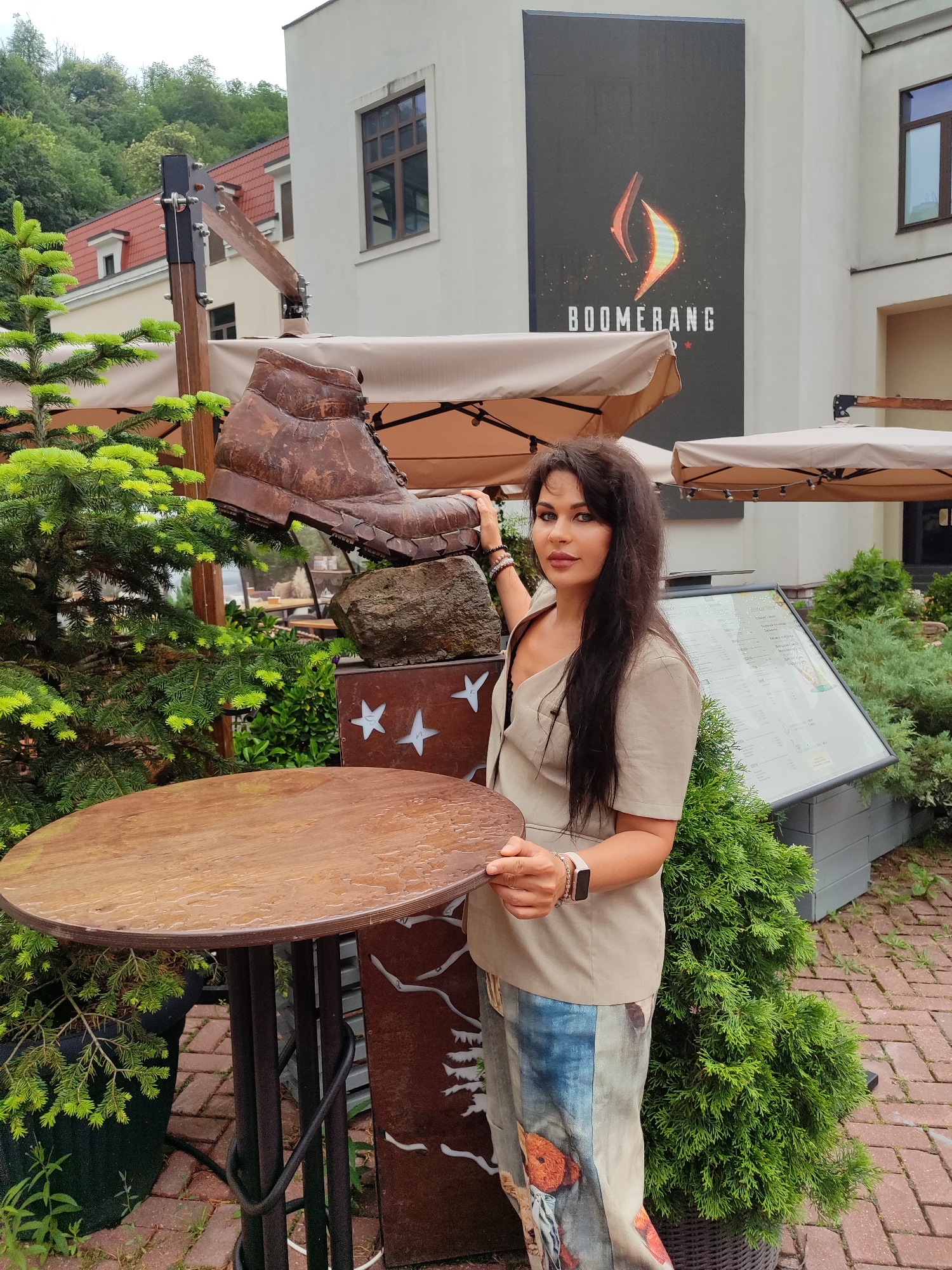 A woman with long, dark hair stands in front of an outdoor seating area. She is wearing a light beige, collared shirt and a long, printed skirt. She is standing near a large, rusty metal sculpture of a boot resting on a rock. To her left, a small evergreen tree stands next to a round wooden table. Behind her, a large screen displays a logo with the word "BOOMERANG" and an image resembling flames. The background shows a building with red-tiled roofs and green trees. The scene appears to be outdoors in an urban or resort setting.