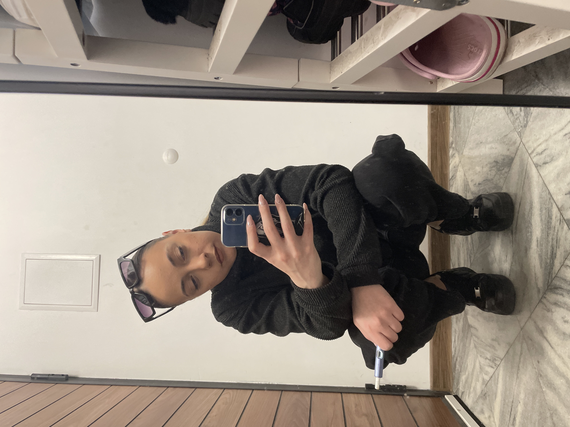 A selfie-style image shows a person squatting indoors, reflected in a mirror. The person is wearing dark clothing, including a sweater, pants, and boots. The person holds a phone up, presumably taking the photo. In their other hand, they are holding a blue object that may be an electronic device or tool. Behind the person, there's a white wall with a recessed square shape. Above the mirror, there's a white storage unit with multiple shelves holding various items, including footwear (like a pink Croc). The floor is light-colored with a marble-like pattern near the right of the image and brown planks on the left edge. It is not possible to determine the location in Sofia, Bulgaria, from this image.