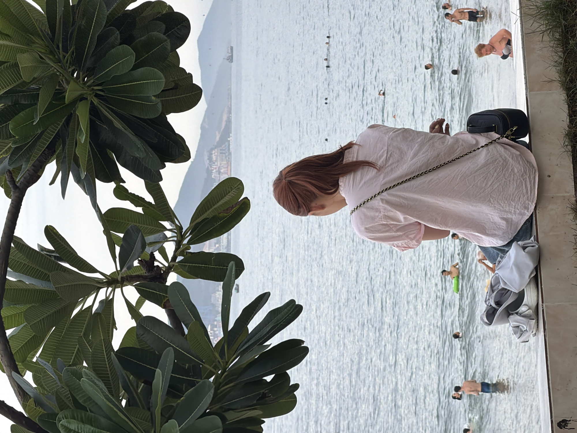 A woman with reddish-brown hair tied in a ponytail, wearing a light pink collared shirt and blue jeans, sits on a white ledge overlooking the ocean. She has a black crossbody bag slung over her shoulder. In front of her on the ledge are a pair of white Crocs and a light gray garment. The ocean is calm with several people swimming and wading in the water. In the distance, a cityscape with mountains and buildings can be seen under a cloudy sky. The foreground is partially obscured by the large, green leaves of a tree. This scene is likely located on the coast of Nha Trang, Vietnam, given the tropical vegetation and coastal landscape.