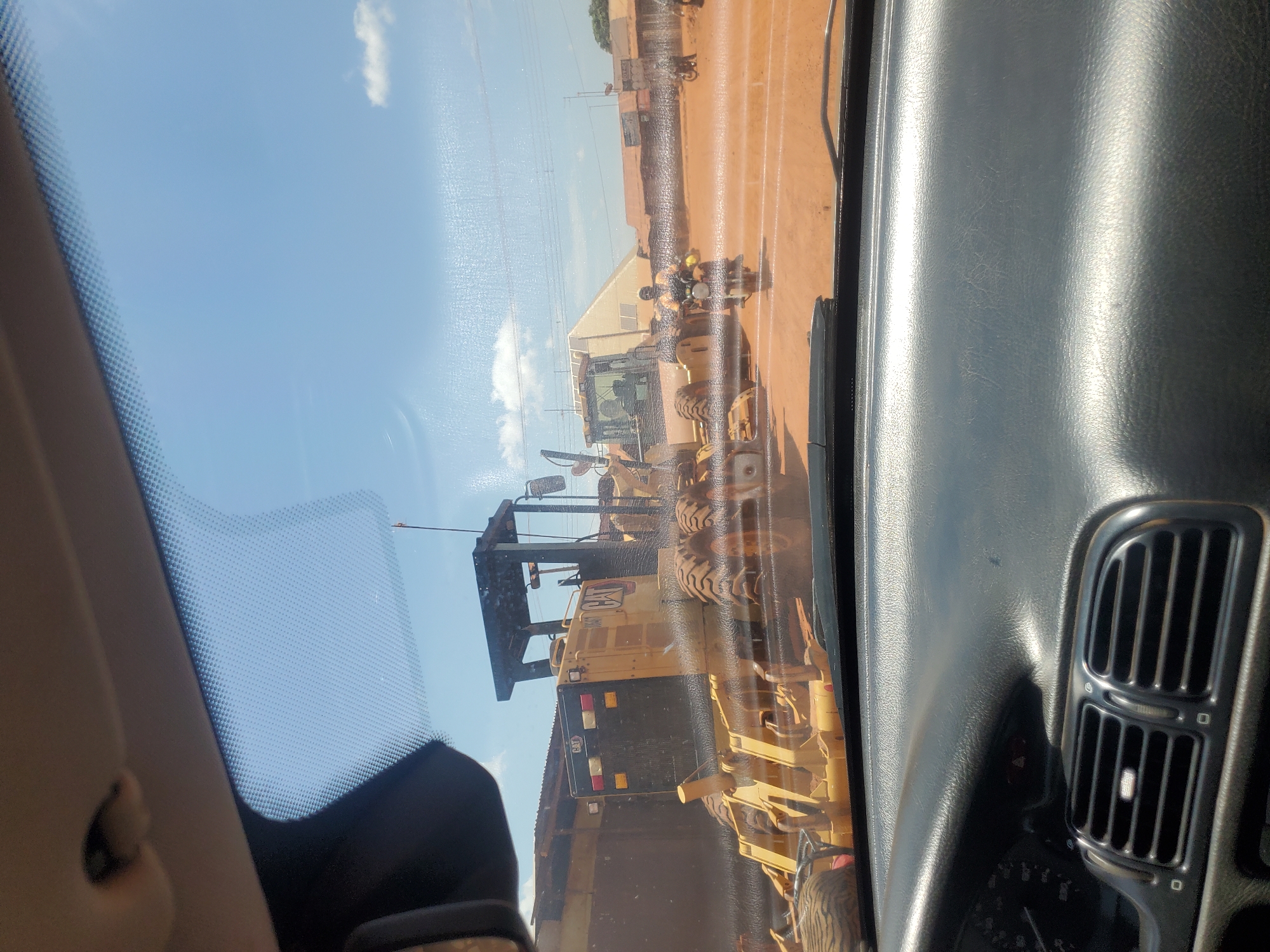The image presents a view from inside a vehicle in Yelwa, Nigeria, looking out through a windshield with visible smudges and watermarks. The vehicle's dark gray dashboard occupies the foreground, displaying air vents on the left and a partial view of the instrument cluster. The upper edge of the windshield shows a dotted frit band, and a section of the vehicle's light-colored headliner is visible at the top.
The outdoor scene is dominated by heavy construction equipment on a reddish-brown dirt surface, indicative of an unpaved road or construction site, under a clear blue sky with scattered white clouds. A prominent yellow Caterpillar (CAT) motor grader is positioned in the mid-ground, observed from its rear-left, with its large tires, engine compartment, and open operator's structure clearly visible. The 'CAT' logo is discernible on its side. To the right and slightly behind this grader, another yellow piece of heavy machinery, possibly a second grader, is partially visible.
Several motorcyclists are present on the dirt road. One individual on a motorcycle, wearing a yellow or orange vest, is centrally located to the right of the main grader, moving away from the viewer. Other motorcyclists are visible further in the background to the right. In the distant background, power lines traverse the frame, and a large, light-colored building with a sloped roof stands behind the machinery, alongside other smaller, indistinct structures on the horizon.