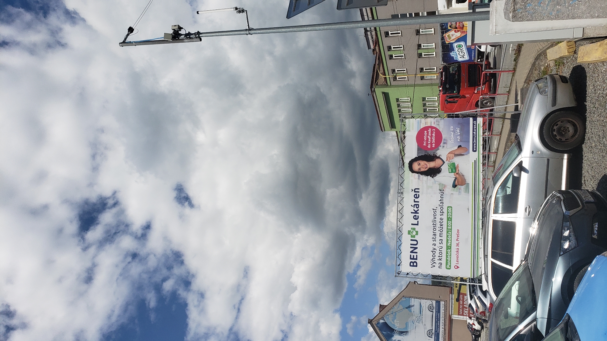 A billboard for BENU Lekáreň is prominently displayed in Prešov, Slovakia. The billboard features a woman in a white lab coat holding two green cards, smiling at the viewer. Text on the billboard includes "BENU Lekáreň" (BENU Pharmacy), "Výhody a starostlivosť, na ktorú sa môžete spoľahnúť!" (Benefits and care you can rely on!), "Pondelok - Nedeľa | 8:00 - 20:00" (Monday - Sunday | 8:00 - 20:00), and "Levočská 36, Prešov" (Levočská 36, Prešov). The billboard also includes the website "www.benulekaren.sk". To the right of the woman, there is text that reads "Pri vstupe do Kauflandu na Sidlisku 3" (Upon entering Kaufland on Sidlisko 3) and "Tešíme sa na Vás" (We look forward to seeing you).
In the foreground, several cars are parked. A silver station wagon is visible in the center, with a dark gray hatchback partially visible on the left and a light blue car in the very left foreground. Behind the BENU Lekáreň billboard, a green building with multiple windows is visible, alongside a red truck and other vehicles. The sky is filled with dramatic, dark gray clouds, suggesting an impending storm, contrasting with patches of blue sky. A utility pole with various electronic equipment is also visible on the right side of the image.
