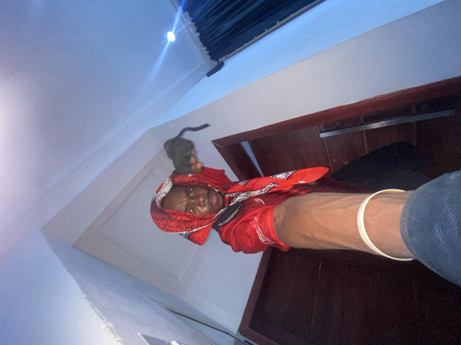 An adult male individual is depicted in an indoor setting within the city of Joma, Ghana. The subject, with dark skin, stands positioned from a low-angle perspective, extending his left arm directly towards the camera, revealing prominent veins and a light-colored wristband. His head is covered by a red and black patterned fabric, creating a hood-like appearance over his short, dark hair. He wears a red short-sleeved top, possibly a jersey, and a black strap bearing "ULTRASPOR" text is visible around his neck. His right hand, covered by a black glove, is clenched into a fist and held near his head, with a black strap hanging from it. The subject maintains an intense, direct gaze into the camera. The background consists of white walls and ceiling, with a distinct ceiling corner visible. A dark wooden doorway or passage is partially obscured behind the subject, and dark curtains are visible in the upper right background. An artificial light source casts a bright, bluish flare on the upper right ceiling.