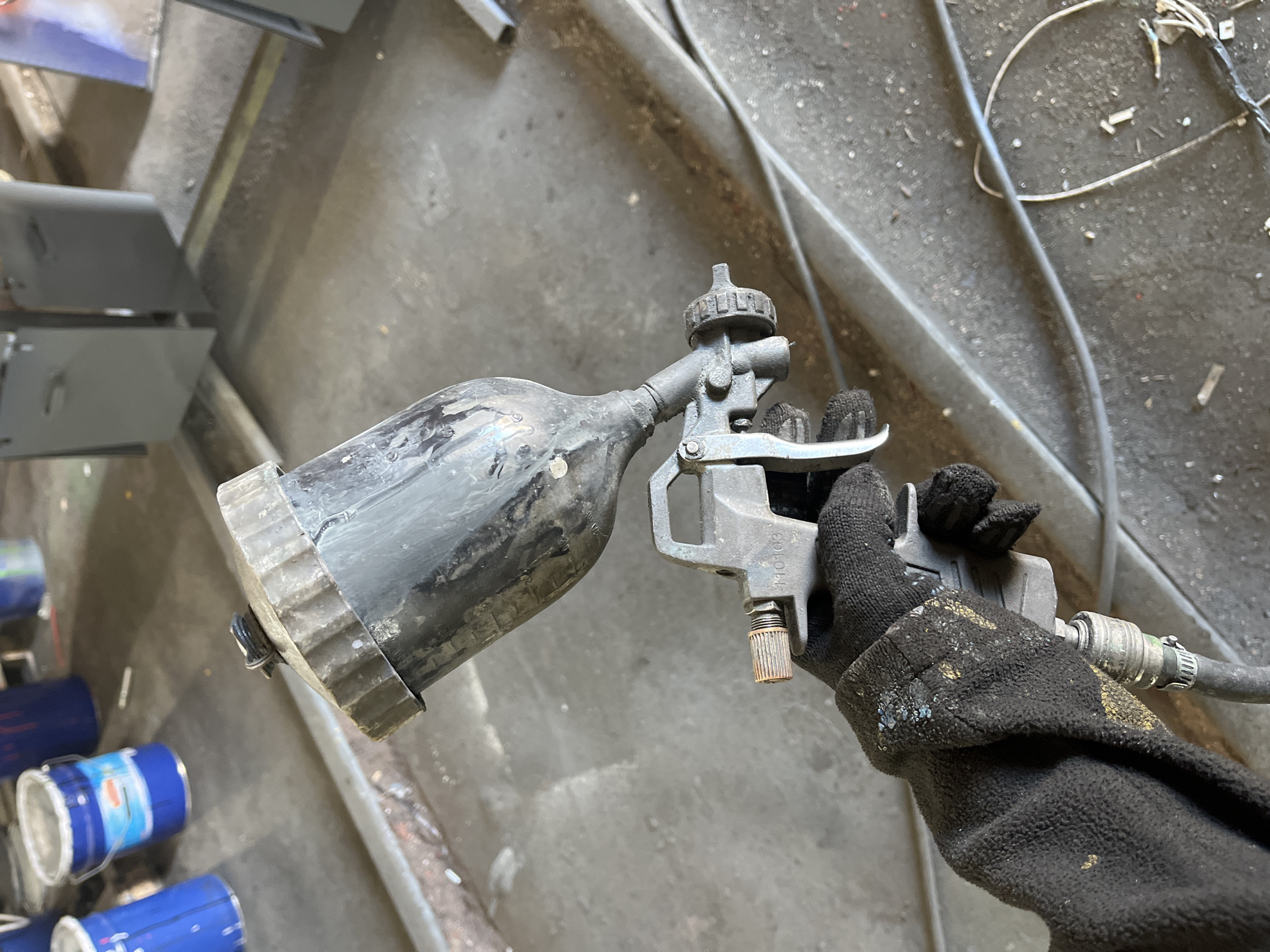 A gloved hand holds a paint spray gun in what appears to be an industrial workshop or painting area in Chaikovsky, Russia. The perspective is top-down, looking at the hand and spray gun, with a dirty concrete floor and various equipment in the background.
The primary subject is a person's right hand, clad in a heavily used, black fabric work glove, possibly stained with gold or yellow paint and other debris. The hand firmly grips the handle of a gravity-feed paint spray gun, which also shows significant signs of wear and use, with dried paint splatters and dirt covering its dark grey or black surface. The paint cup, mounted on top of the gun, is equally dirty. A metal clamp connects a hose, likely for compressed air, to the base of the gun's handle. A rusted adjustment knob is visible on the gun's lower body. The numbers "110103" are faintly visible on the metallic body of the spray gun near the trigger mechanism.
In the background, a grimy concrete floor is visible, strewn with wires, cables, and various small pieces of debris. To the left, several blue paint cans are stacked or placed, suggesting the nature of the work being performed. Further back, what appear to be grey metal panels or parts are stacked, possibly awaiting or having just received a coat of paint. The overall setting conveys a working environment focused on painting or finishing metal components.