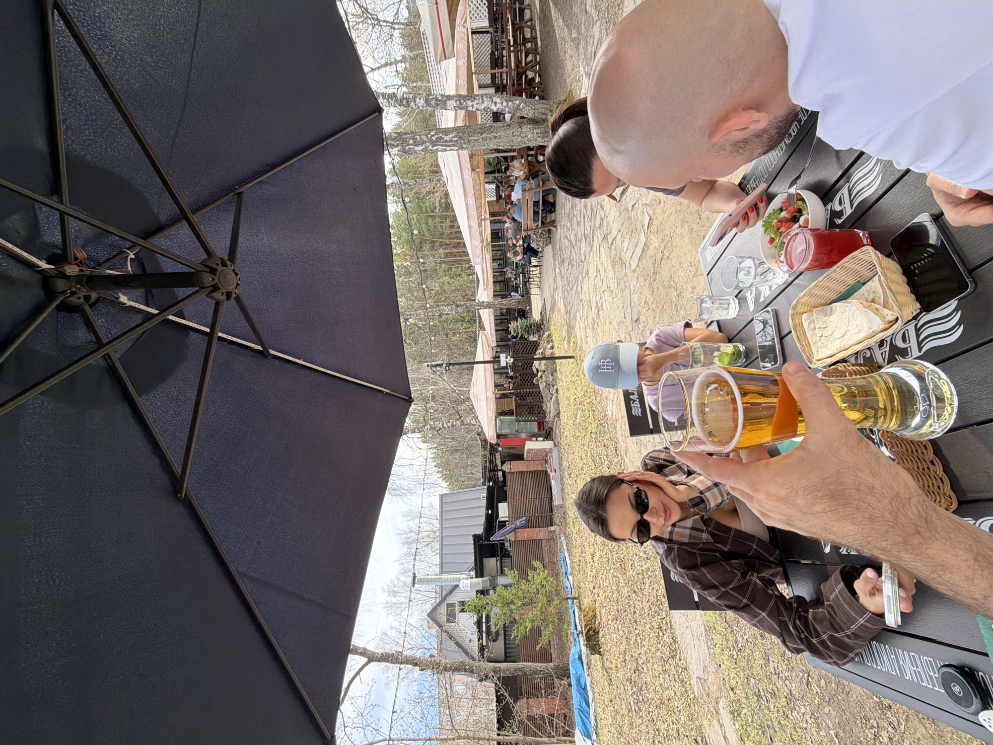 A casual scene unfolds at what appears to be an outdoor dining area in Krasnoyarsk, Russia, identifiable by Cyrillic lettering visible on a table accessory. The foreground is dominated by a large, black patio umbrella, obscuring part of the view. Around a black table, several individuals are gathered.
One person, wearing a brown plaid jacket and sunglasses, is visible to the camera's left. Directly across from this individual is another person wearing a light purple shirt and a blue hat. To the right, a bald man in a white shirt is visible, partially out of frame, along with salad and bread on the table. The hand and arm of another individual, raising a beer glass, enter the frame from the bottom right.
Several drinks and a small basket of flatbread are placed on the table alongside mobile phones and other accessories. The dining area is situated outside, with other tables and customers visible in the background. Trees and buildings are also visible in the background, suggesting a more rustic, outdoor setting. The overall impression is a group of people enjoying a meal and drinks in an outdoor setting.