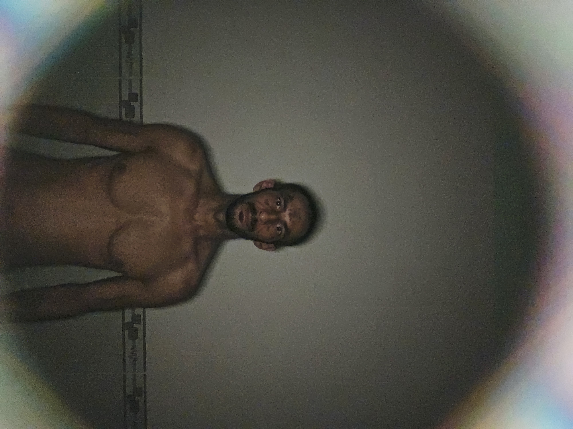 The image displays a shirtless man, positioned horizontally within the frame, looking directly at the camera. His body is oriented with his head towards the upper right and his torso extending towards the lower left. He has dark skin, a beard, and a neutral to slightly surprised expression, characterized by wide eyes and a slightly open mouth.
The background consists primarily of a plain, dark gray wall. On the left side, a narrow section of a tiled wall is visible, showing a subtle pattern. The lighting is dim, resulting in a noticeable grainy texture across the entire image. A prominent lens flare effect is present around the perimeter of the frame, creating a colorful, rainbow-like glow with discernible hues of purple, blue, green, and red, particularly along the top and right edges. This effect suggests a direct light source interacting with the camera lens. No text is visible in the image. The indoor setting and artificial lighting prevent determination of the time of day.