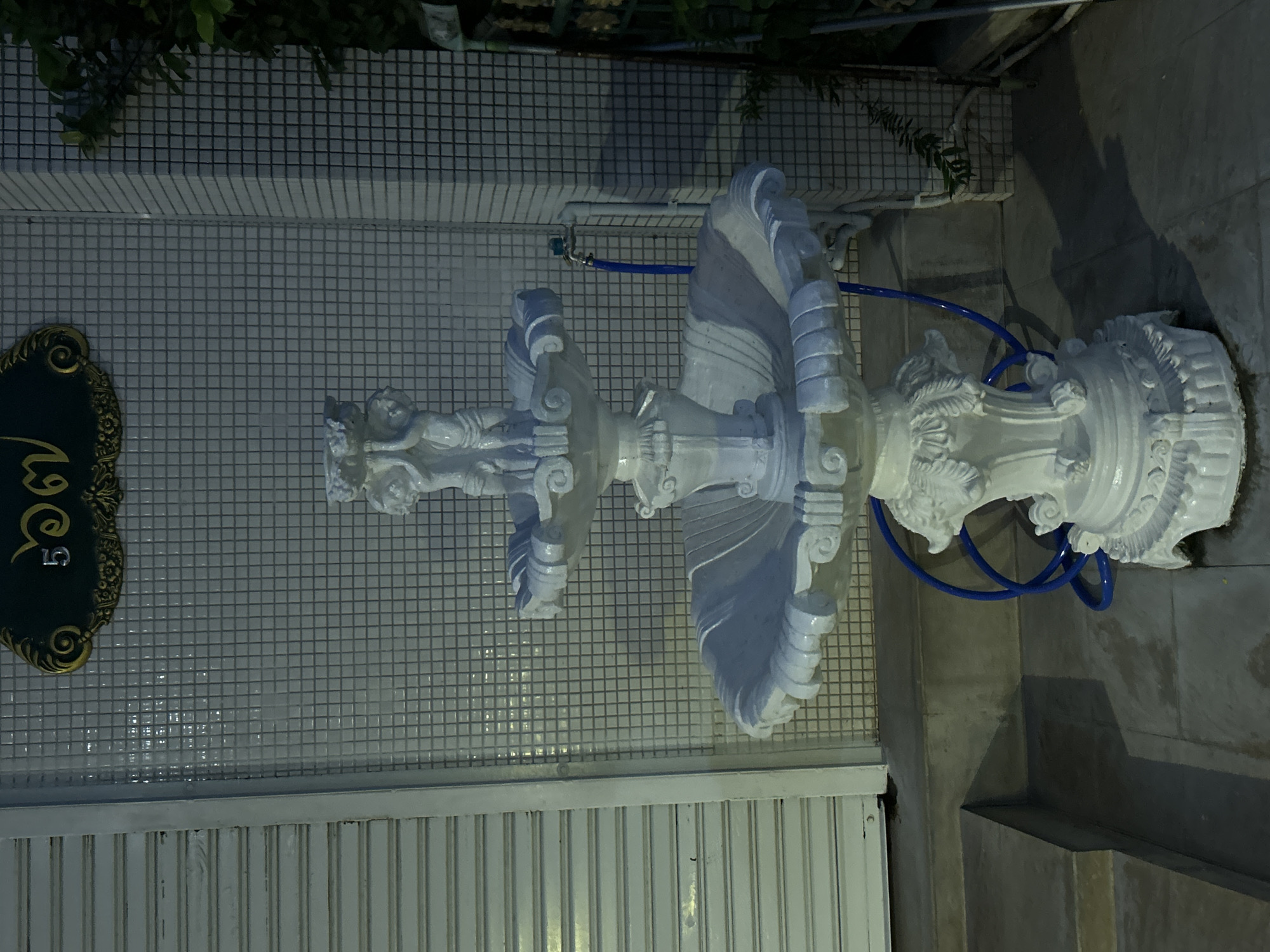 A white, multi-tiered fountain with decorative cherubic figures and ornate bases stands in front of a tiled wall in Phuket City Municipality, Thailand. A blue hose is coiled on the ground near the fountain's base, and a faucet is visible on the wall behind it. The scene is illuminated by artificial light, casting shadows on the tiled floor and a set of concrete stairs to the left. A closed metal shutter is also visible to the left of the fountain. A dark, ornate sign with Thai numerals "5 ๑ 6" is mounted on the tiled wall above the fountain. Green foliage is present to the right of the fountain.