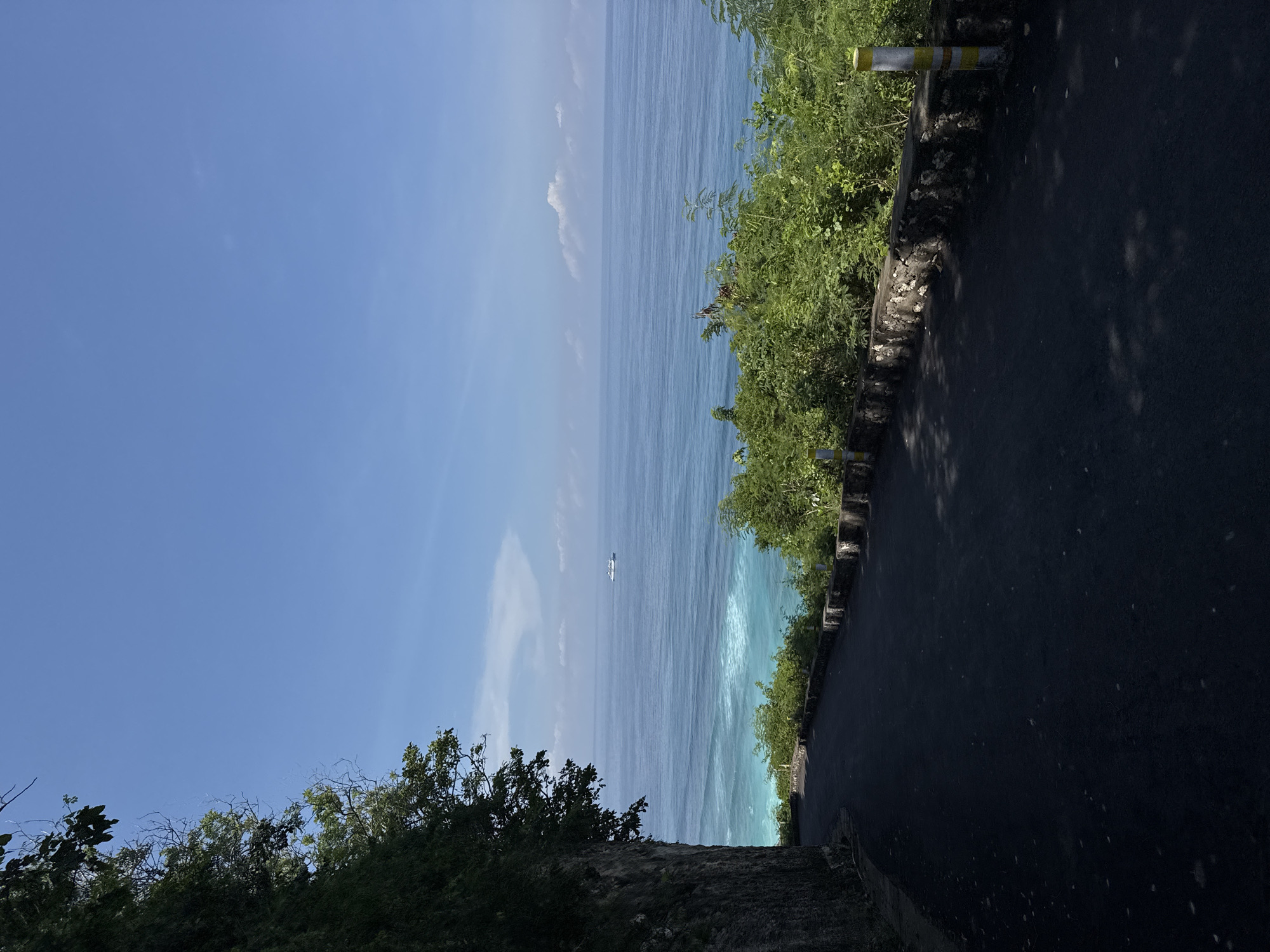 The image shows a seaside road scene, likely in Kuta Selatan, Indonesia. The viewpoint is from a road that runs along the top of a cliff. To the left, the bright blue sky transitions into the blue ocean. A small white object, possibly a boat, is visible in the water. On the right, the dark grey road edge has green shrubs and vegetation. A white and yellow striped post are placed along the road edge. Trees obscure the view on the lower-left. The road surface is dark due to shadows. The overall environment suggests a coastal road on a sunny day.
