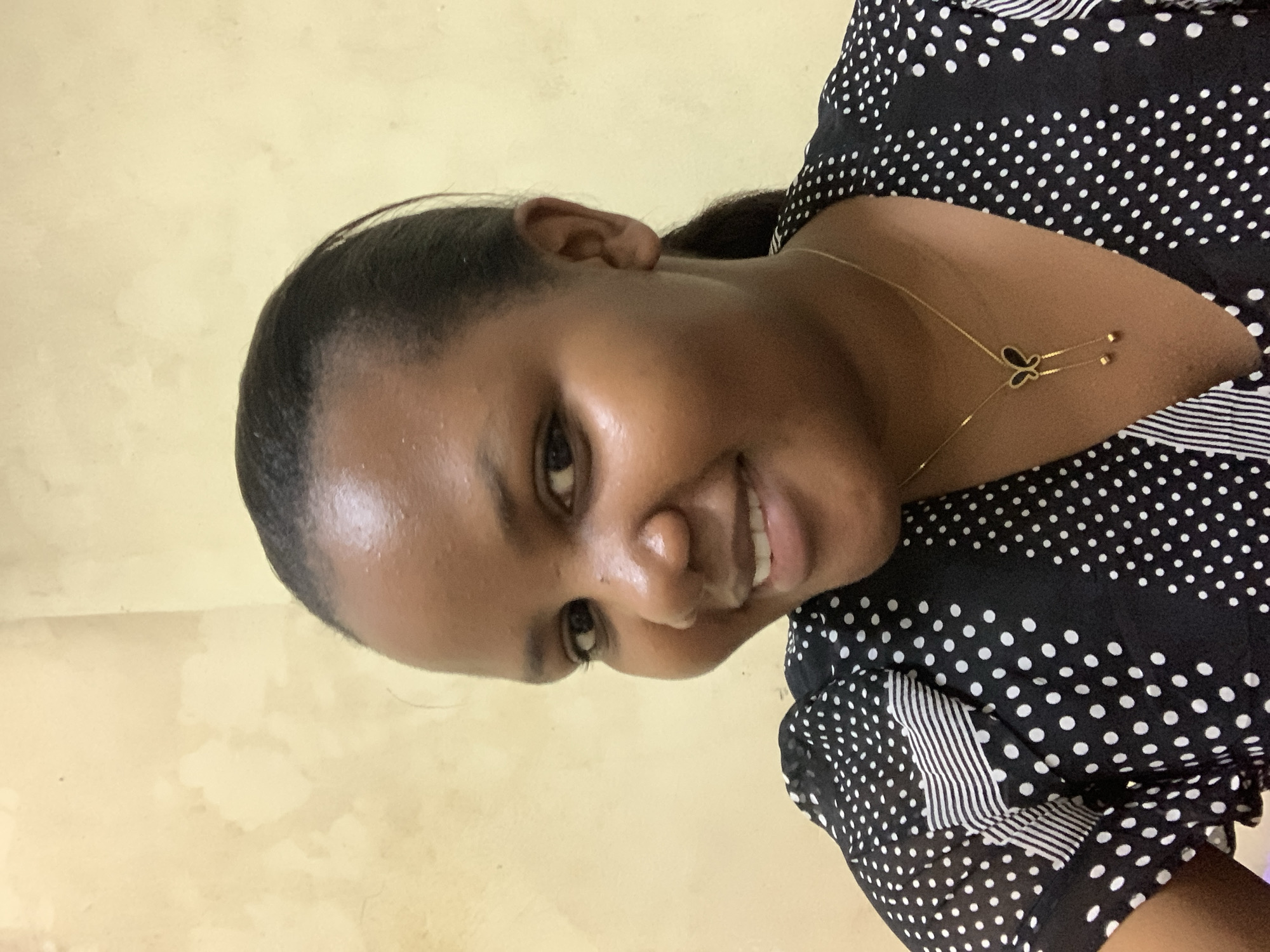 A woman with dark skin and black hair is smiling at the camera. She is wearing a black and white polka dot dress and a gold necklace with a butterfly pendant. The background is a yellowish-tan wall. There is no information about the location being in Dar es-Salaam, Tanzania.
