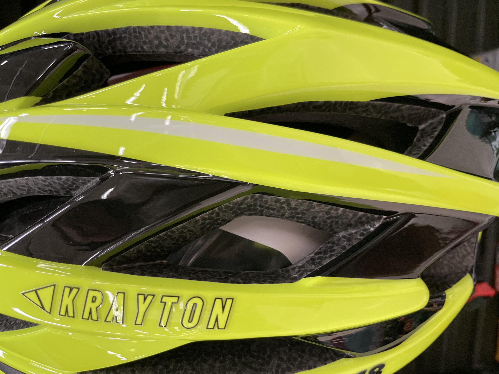 The image shows a close-up of a bright yellow bicycle helmet. The helmet has a glossy finish and black accents, with a prominent white stripe on one side. The brand name "KRAYTON" is visible in raised, silver lettering on the left side of the helmet, with a small triangle above the "K." The helmet's interior padding is visible in some of the vents, appearing to be grey and textured. The lighting suggests an indoor setting, possibly a retail display or a storage area. The image does not provide any information or visual cues to determine a specific location in the city of Málaga, Spain.