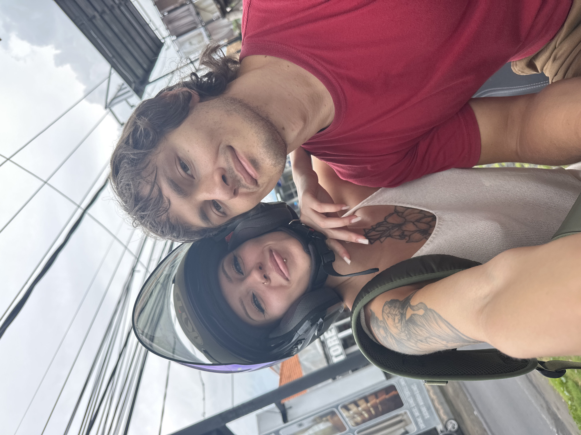 The image shows two individuals, a man and a woman, in Munggu, Indonesia, both appearing to be of Caucasian descent. The woman is wearing a black motorcycle helmet with a clear visor, and a light-colored top. Her chest features a floral tattoo. A large tattoo is visible on her arm as well. The man is wearing a red t-shirt. They are close to each other, likely on a motorcycle. Background elements include overhead power lines, buildings, and potentially a vehicle.