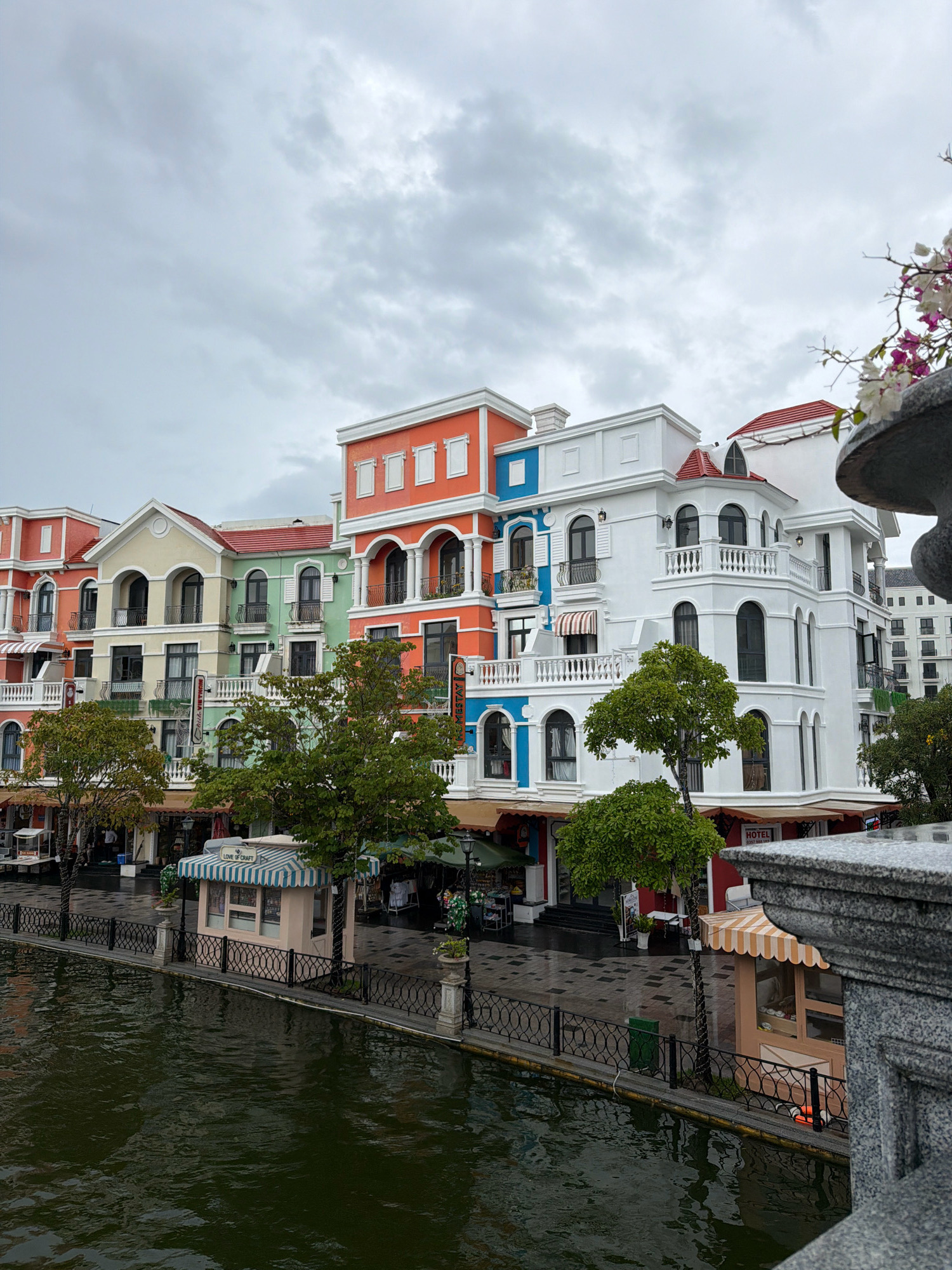 The image depicts a picturesque waterfront promenade in Phu Quoc, Vietnam, likely within the Sun World Mediterranean village. A row of colorful, multi-story buildings with European-inspired architecture lines the promenade. These buildings feature balconies with ornate railings, arched windows, and a variety of vibrant facades in orange, yellow, green, and white. Some buildings have red tiled roofs.
Along the promenade, there are several small kiosks or shops, one of which has a striped awning and a sign reading "LOVE OF CRAFT." Another kiosk has an orange and white striped awning. Trees with lush green foliage are interspersed along the waterfront, providing shade. A black metal fence separates the promenade from the dark, reflective water of a canal or river. The ground of the promenade appears to be paved with stone or tile.
In the background, more buildings are visible, suggesting a developed commercial or residential area. The sky is overcast with heavy, gray clouds, indicating a potentially rainy or gloomy day. A stone balustrade and a large, gray, sculpted object, possibly a planter or decorative element, are visible in the foreground on the right, framing the scene. There are no prominent individuals clearly visible, but a few figures can be discerned in the background near the shops, possibly pedestrians or staff. The overall atmosphere is one of a charming, European-style tourist destination.