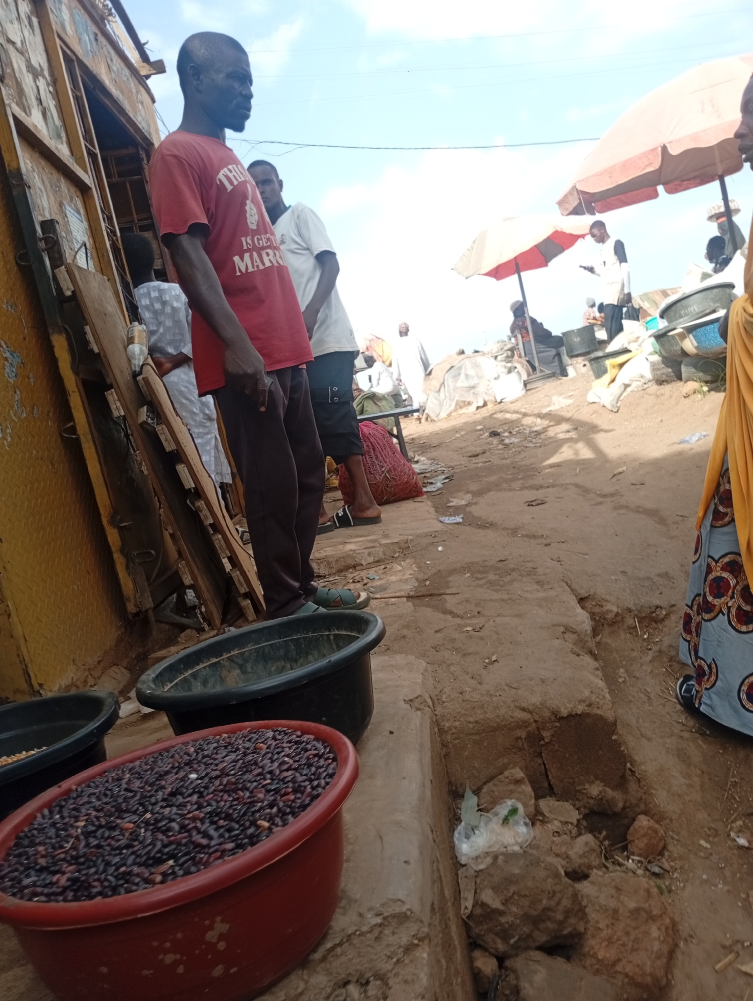 A medium shot captures an outdoor marketplace scene with an emphasis on a man in the foreground. The man, appearing to be in his late 40s to early 50s, stands with his left side to the camera, wearing a red t-shirt with white lettering that partially reads "THIS IS GETTING MARRIED." He is also wearing dark brown trousers and green sandals. To his right, another man in a white t-shirt and dark trousers is partially visible. In the lower left of the frame, a large red tub is filled with dark, small, bean-like items. Behind this tub, a dark green tub is also visible, with a smaller dark tub to its left, containing a yellow granular substance. The ground is dusty and uneven, with several rocks and debris scattered around. In the background, there are several market stalls, some with umbrellas, and various people milling about. The sky is visible with some clouds, suggesting an outdoor, daytime setting. The overall atmosphere suggests a busy, local market in Rafin Par, Nigeria.