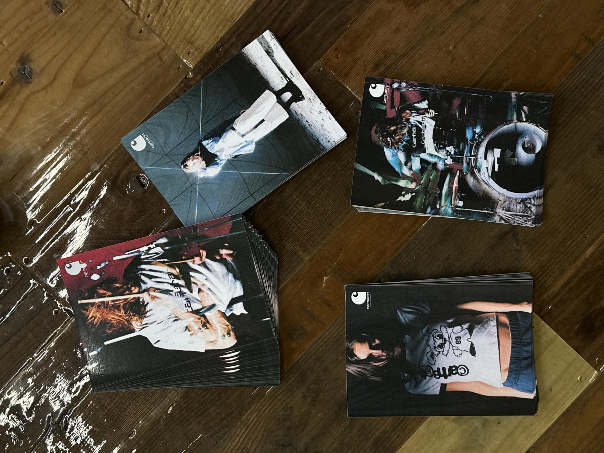 The scene depicts a wooden surface with four stacks of promotional cards. Each card features different images of people wearing Carhartt clothing. The top left card shows a person in white pants and a light blue shirt. The top right card features a person obscured in a dark, industrial setting. The bottom left stack contains a large number of cards with an image of a person with reddish-brown hair, holding drumsticks, wearing a white shirt and dark pants. The bottom right card shows a person wearing a white Carhartt t-shirt and blue shorts.
The location in Paris, France cannot be determined from the visual information provided.