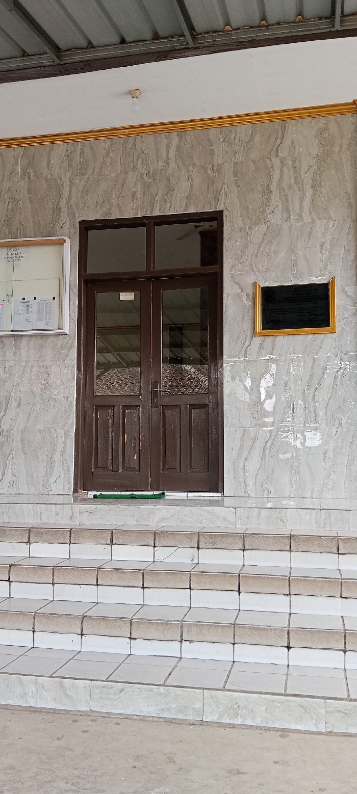 The image shows the front entrance to a building in Kecamatan Kutawaringin, Indonesia. The building is made of white marble tiles with a gold trim. The entrance has a set of double wooden doors with glass panels, and a small green mat in front of them. The doors lead to a hallway. There are five steps leading up to the door. There is a light fixture on the ceiling above the entrance and a small plaque on the wall to the right of the doors. The plaque is gold-framed and contains a black background. There is a bulletin board to the left of the doors with a white background and a black frame. The surrounding environment is a concrete walkway, a light grey in color. There are no people or objects in front of the door.