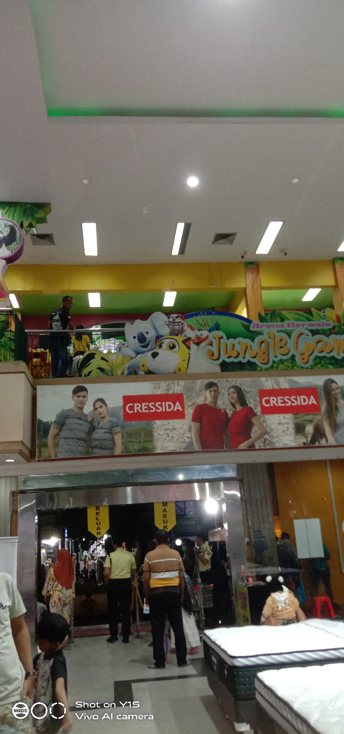 The scene depicts a multi-level indoor commercial environment, specifically within a shopping mall or large department store located in Probolinggo, Indonesia.
**Environment:** The overhead ceiling is white, featuring multiple recessed circular lights and linear fluorescent strip lights. A perimeter dropped ceiling section is illuminated with green accent lighting. Air conditioning vents are visible. An upper level or balcony, defined by a yellow painted wall and green leaf-motif decorations, features a prominent sign for "Jungle Game" with "Arena Bermain" (Play Area) above it. This area displays several large, colorful cartoon animal figures, including a koala, a tiger, and a bird-like creature. A metal railing separates this upper level from the lower viewing area. Below this, a large horizontal banner advertises the brand "CRESSIDA," featuring models in casual attire: one pair in striped gray t-shirts and another in solid red t-shirts. The ground level includes an entrance/exit point delineated by two yellow hanging banners, "KELUAR" (Exit) on the left and "MASUK" (Enter) on the right. In the immediate foreground to the right, two mattresses with white tops and dark bases are on display.
**People:** Approximately 12-15 individuals are visible across both levels. On the upper level, four to five people are present, including a man in a dark shirt and backpack near the railing, and others further back. On the ground level, a person in a light t-shirt, partially visible on the far left, holds the hand of a young child in a dark patterned shirt. A woman in a floral dress and orange head covering walks away from the entrance. Two men, viewed from behind, stand near the entrance: one in a light yellow shirt, the other in a brown and yellow striped shirt. Another individual, possibly a security guard, is present near a metal shopping cart to the right of the entrance, wearing a light uniform shirt, dark pants, and a face mask. A small child, wearing a beige top with black ear-like appliques, is seated on one of the displayed mattresses on the far right.
**Actions/Interactions:** Individuals are observed walking, standing, and navigating the commercial space. A child is being accompanied by an adult, and another child is stationary on a retail display. No direct interactions between individuals are clearly discernible.