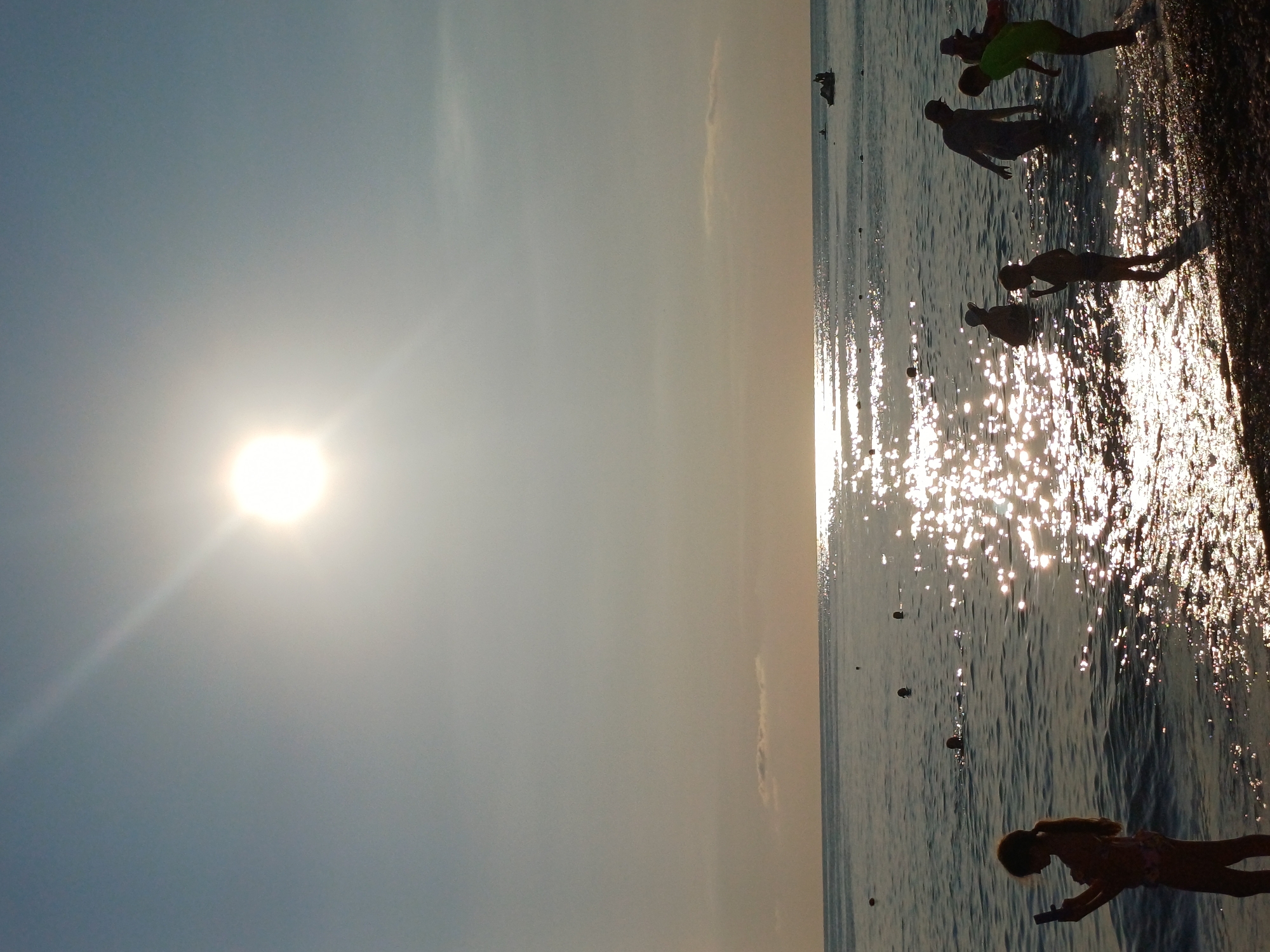 The image depicts a sunny day at a beach, likely in Sochi, Russia, judging by the presence of a water jet ski in the background. The sun is high in the sky, creating a bright glare on the water. Several people are visible, including a young girl standing on the shore holding a phone, a group of children and adults wading in the shallows, and some individuals swimming further out. The water is calm with gentle waves lapping at the shore. The beach itself appears to be composed of pebbles. The overall atmosphere is one of a leisurely day spent at the seaside.