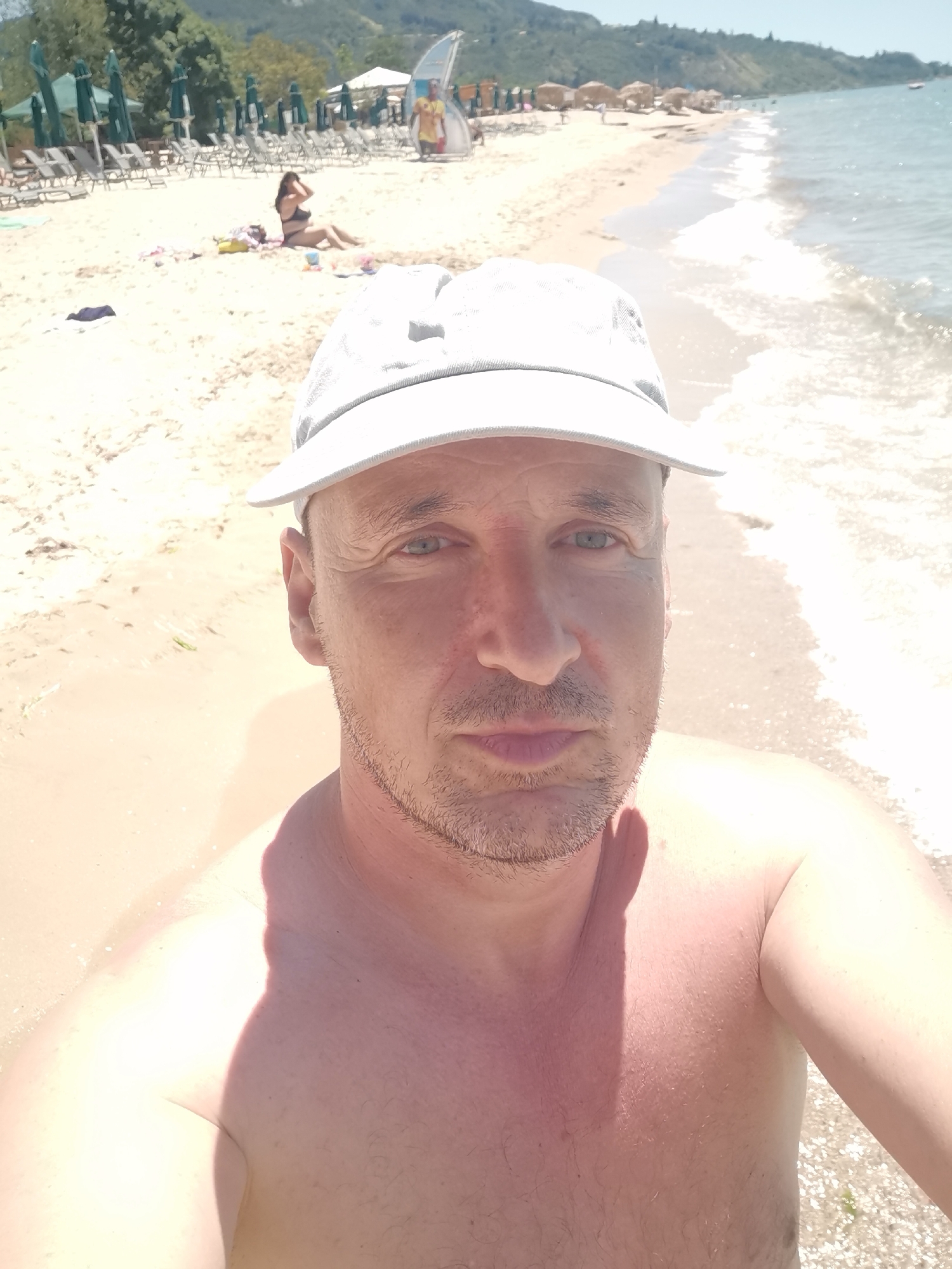 A man wearing a white cap takes a selfie on a sandy beach. He is shirtless, showing his shoulders and chest. The beach is lined with lounge chairs and umbrellas in the background. The sea is calm and blue. The man appears to be in his 40s, with short brown hair and a beard. He is looking directly at the camera with a neutral expression. The location is Kranevo, Bulgaria.