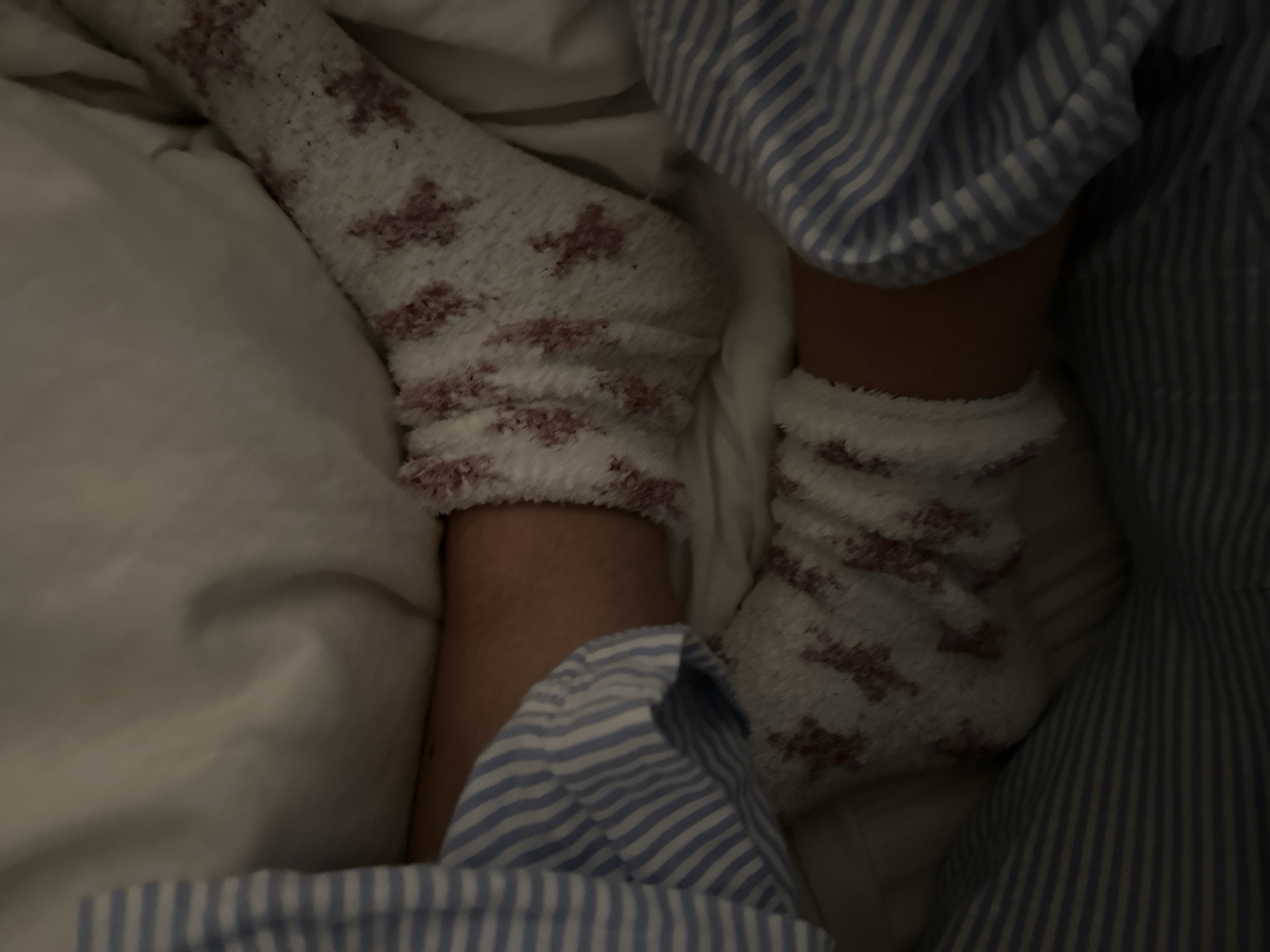 The scene is located in a dimly lit indoor environment, specifically depicting a bed in Wealden, United Kingdom. Two human lower legs/feet are visible, resting. Both feet are clad in fluffy white socks featuring a repeating pattern of reddish-brown/maroon bow-like or cross-like motifs. Portions of a blue and white striped fabric, consistent with sleepwear or bedding, are present on both the left and right sides of the frame, partially covering the legs and blending with the surrounding light-colored bedding material, likely a duvet or sheet. No faces or specific individuals are identifiable; the focus is on the feet and immediate bedding. The overall impression is one of repose.