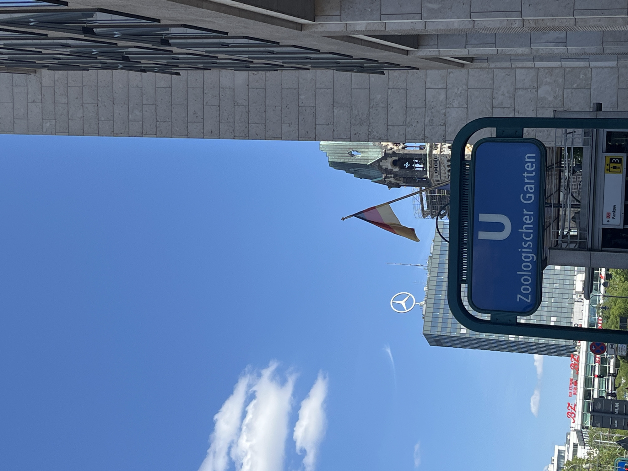 The image shows a bright, sunny day in Berlin, Germany, with a clear blue sky and a few wispy white clouds. The perspective is from below, looking upwards, and the image is rotated 90 degrees clockwise.
In the foreground, a blue sign with white text reads "U Zoologischer Garten". This indicates the Zoologischer Garten U-Bahn (subway) station, a well-known location in Berlin. The sign is mounted on a dark green metal frame.
Behind the sign, several modern glass and concrete buildings rise into the sky. One prominent building features a large white Mercedes-Benz logo on its facade. Another building has a distinctive architectural detail, possibly a historical tower or spire, with a green roof and intricate stonework. A German flag, with horizontal stripes of black, red, and gold, is visible on a flagpole.
The scene appears to be an urban environment, likely a busy street or plaza near the Zoologischer Garten station. The bright sunlight and clear sky suggest it's daytime, possibly late morning or early afternoon. There are no people clearly visible in the frame, but the presence of the U-Bahn sign and the buildings implies a public space. The overall impression is one of a vibrant, modern city under a pleasant sky.