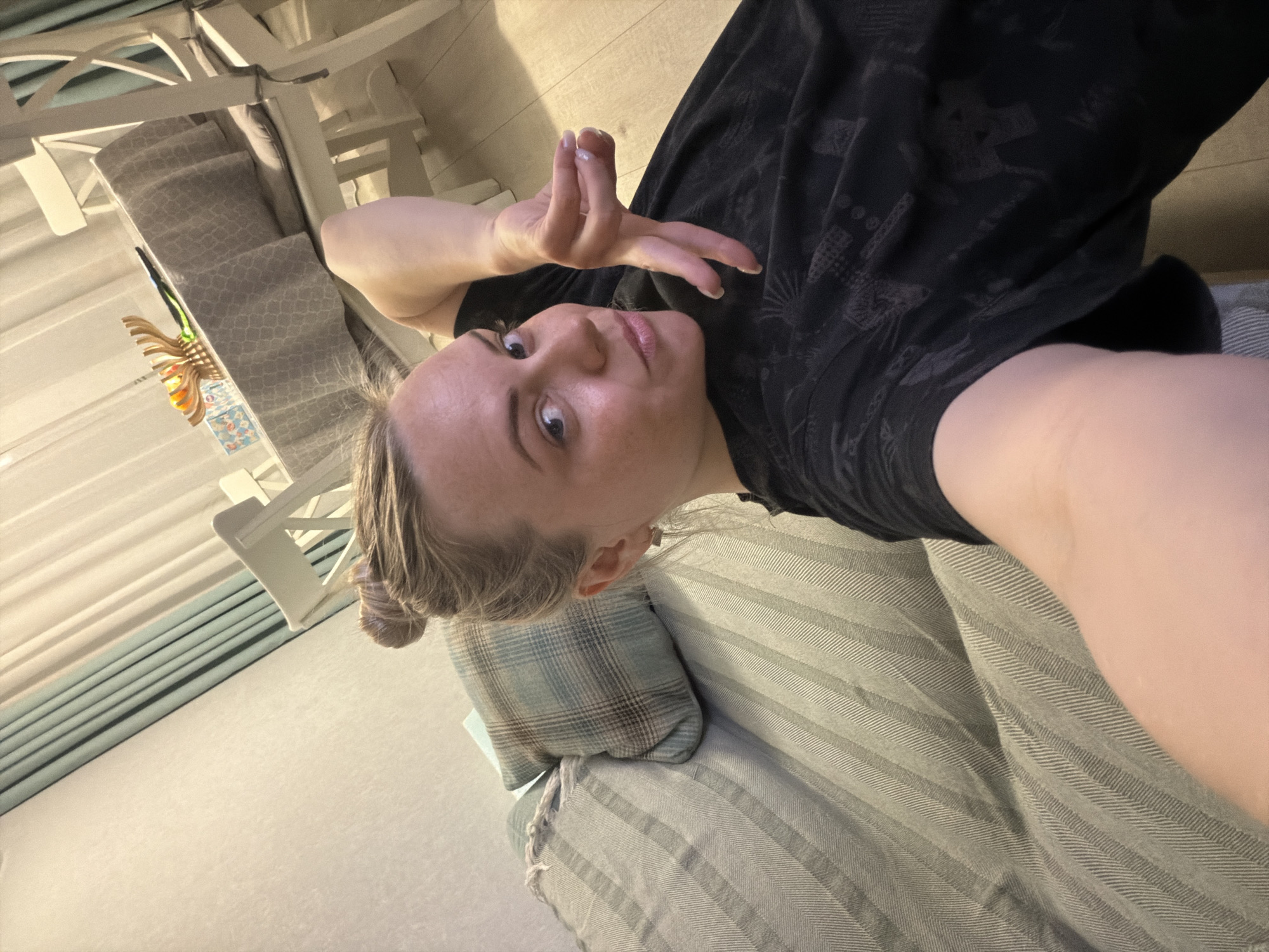 A selfie photograph shows a woman lying on a striped cushion with a plaid pillow behind her head. She is wearing a black t-shirt with printed designs. Her hair is blonde and pulled back into a bun. She is holding up her right hand with her fingers slightly splayed. A white chair and a decorative wall hanging are visible in the background. The location cannot be determined from the image, specifically in relation to the city of Votkinsk, Russia.