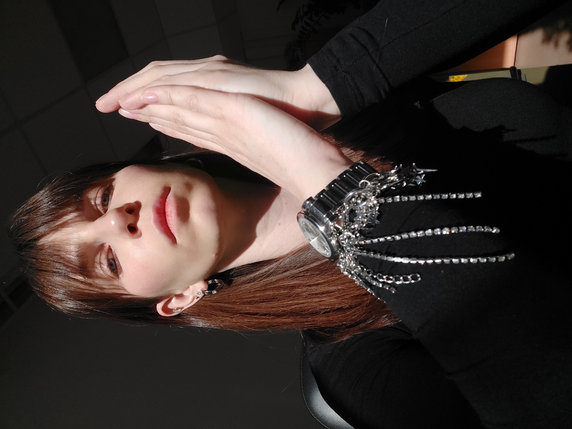 A woman is pictured from a low angle, appearing to be in an indoor setting. She has light skin, brown hair with bangs, and is wearing a black top. She has a watch on her wrist, decorated with chains and rhinestones. Her hands are posed together in the upper part of the frame. The lighting is dramatic, with one side of her face illuminated and the other in shadow. Based on the details provided in the prompt, the location may be in the city of Chaikovsky, Russia, but there is no direct evidence of this visible in the image itself.