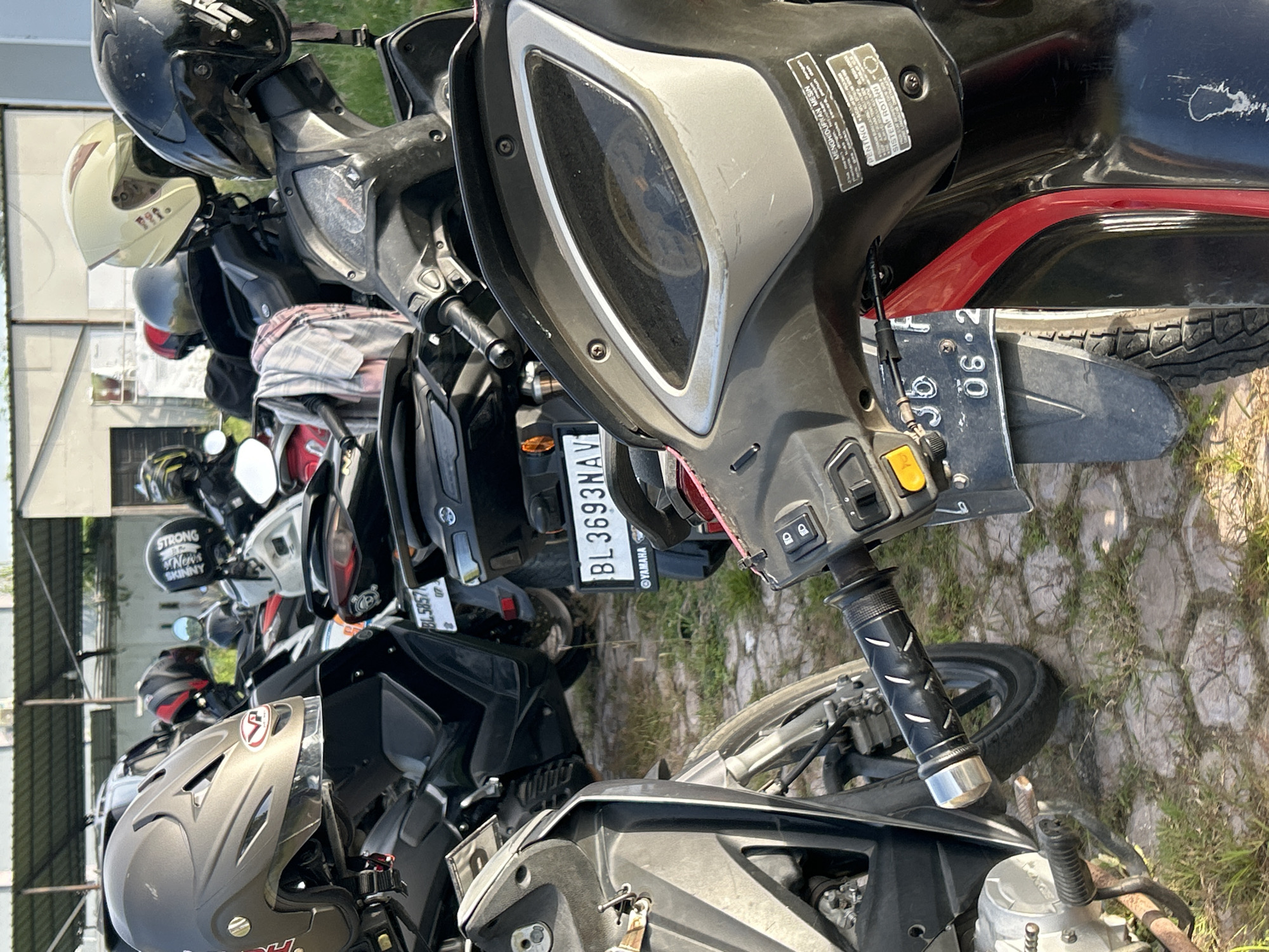 The image shows a row of parked motorcycles. The foreground features a black and gray motorcycle with a license plate obscured by the motorcycle in front of it. The handlebars and front wheel are visible, resting on the ground covered with patchy grass and cracked paving. Other motorcycles of various colors and models are lined up behind, some with helmets perched on them. The license plates of a few motorcycles are partially visible, showing registrations like "BL 3693NAV" and "BL5857." The background shows a building and a cloudy sky. Text visible on the motorcycles includes "YAMAHA," "STRONG," "SKINNY," and "News," along with Indonesian words like "MENGHIDUPKAN MESIN" and "PENTING." The overall scene suggests a typical motorcycle parking area in Indonesia.