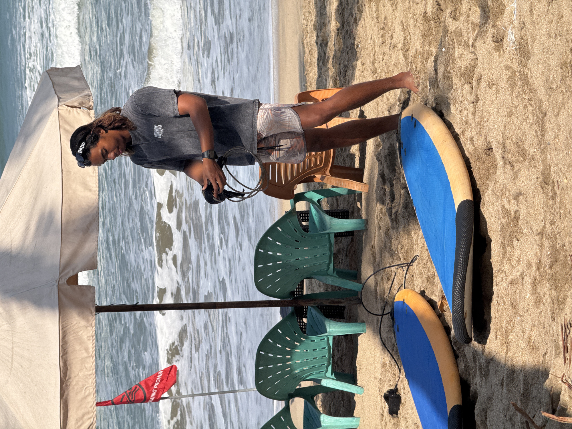 A young man stands on a sandy beach in Kuta, Indonesia, holding a coiled leash for a surfboard. He is wearing a dark grey t-shirt with white lettering and patterned shorts. A black headband with white lettering is on his head, and sunglasses are pushed up onto it. In front of him lie two blue surfboards with yellow and black accents. Behind him, several green plastic chairs and one orange plastic chair are arranged on the sand. A large, light-colored beach umbrella casts a shadow over the scene. To the left, a red flag with white lettering and a symbol is planted in the sand. The ocean, with moderate waves, is visible in the background. The scene suggests a surfing lesson or rental operation.