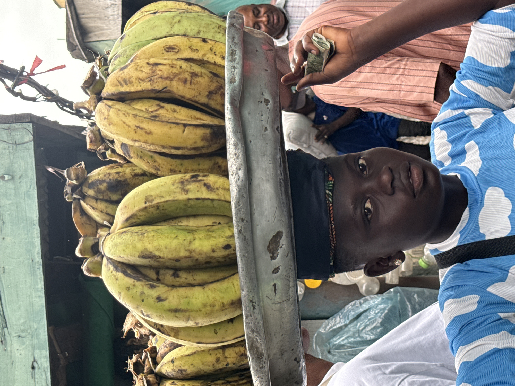 A young person balances a large tray of ripe plantains on their head in a bustling outdoor market scene in Lagos, Nigeria. The plantains, a mix of yellow and green, are clustered in bunches and appear to be for sale. The person is wearing a blue and white patterned shirt and a black headwrap. In the foreground, a hand is shown holding some currency, suggesting a transaction is taking place. Other people are visible in the background, indicating a busy marketplace environment. The overall atmosphere suggests a typical day in a vibrant Nigerian city.
