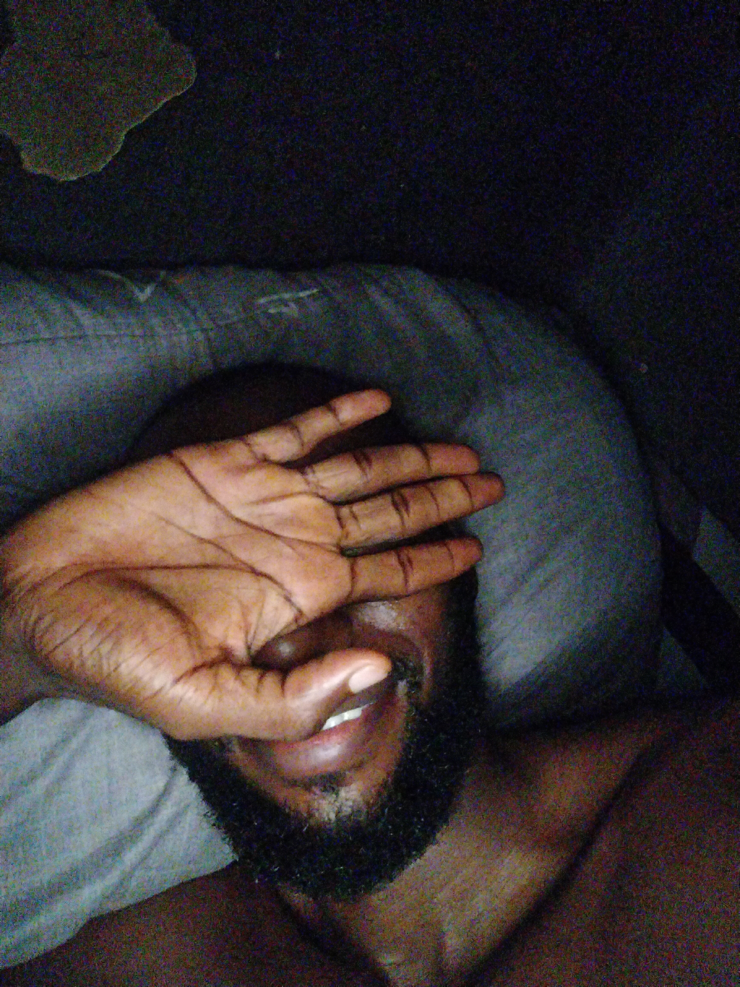 A close-up, dimly lit selfie shows a person's left hand covering their face. The hand is positioned over the eyes and forehead, with fingers spread. A portion of the person's mouth, showing teeth and lips, is visible below the hand, along with a dark beard. The person appears to be lying down, with a pillow visible beneath them. The background is dark and indistinct. The image appears to have been taken indoors, possibly in a bedroom, given the presence of a pillow and bedding. Without further context, it is impossible to determine the specific location within the city of Uyo, Nigeria.