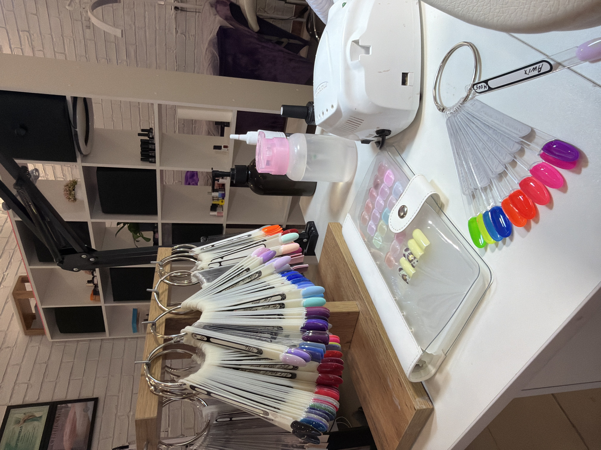 The image shows a nail salon setup. The equipment includes a white airbrush machine, assorted nail polish bottles, and multiple color palettes displaying a range of nail polish colors on white plastic sticks, held together with metal rings. One color palette has "Awix" written on it. A transparent plastic case with various nail decorations is also visible. In the background, a white shelving unit is stocked with storage boxes and beauty products. The salon appears to be in a room with a white brick wall. There is no explicit information about the location in the city of Reshetnikovo, Russia.