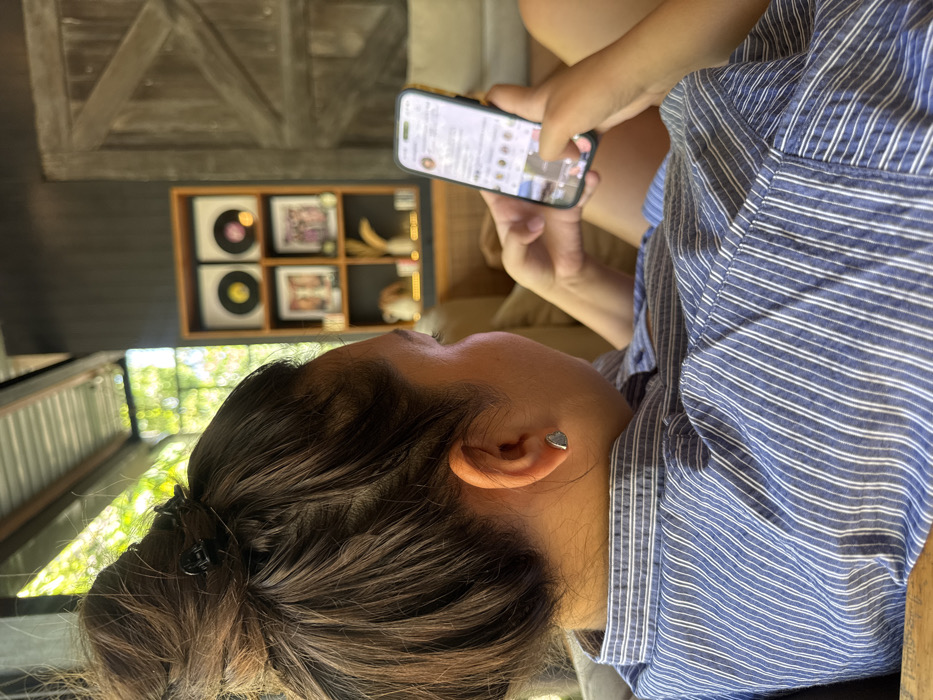 The image captures an individual, seen from behind and the right side, seated in a relaxed position within an establishment in Pecatu, Indonesia. The subject has dark hair pulled into a high, messy bun secured with a black claw clip, and is wearing a blue and white striped button-up shirt. A small, triangular silver earring is visible in their right ear. Both hands are actively engaged with a smartphone, fitted with a yellow case, displaying a social media interface, specifically an Instagram feed. The background features natural light and blurred green foliage overhead, indicating an open or semi-open setting. To the right, a dark paneled wall holds a wooden shelving unit containing two framed vinyl records or circular artworks on the upper shelf, and framed photographs alongside a decorative vase on the lower shelf. Adjacent to this unit is a large, rustic wooden door with diagonal planking. The environment suggests a casual cafe or lounge setting, incorporating dark wood elements and light-colored soft furnishings.