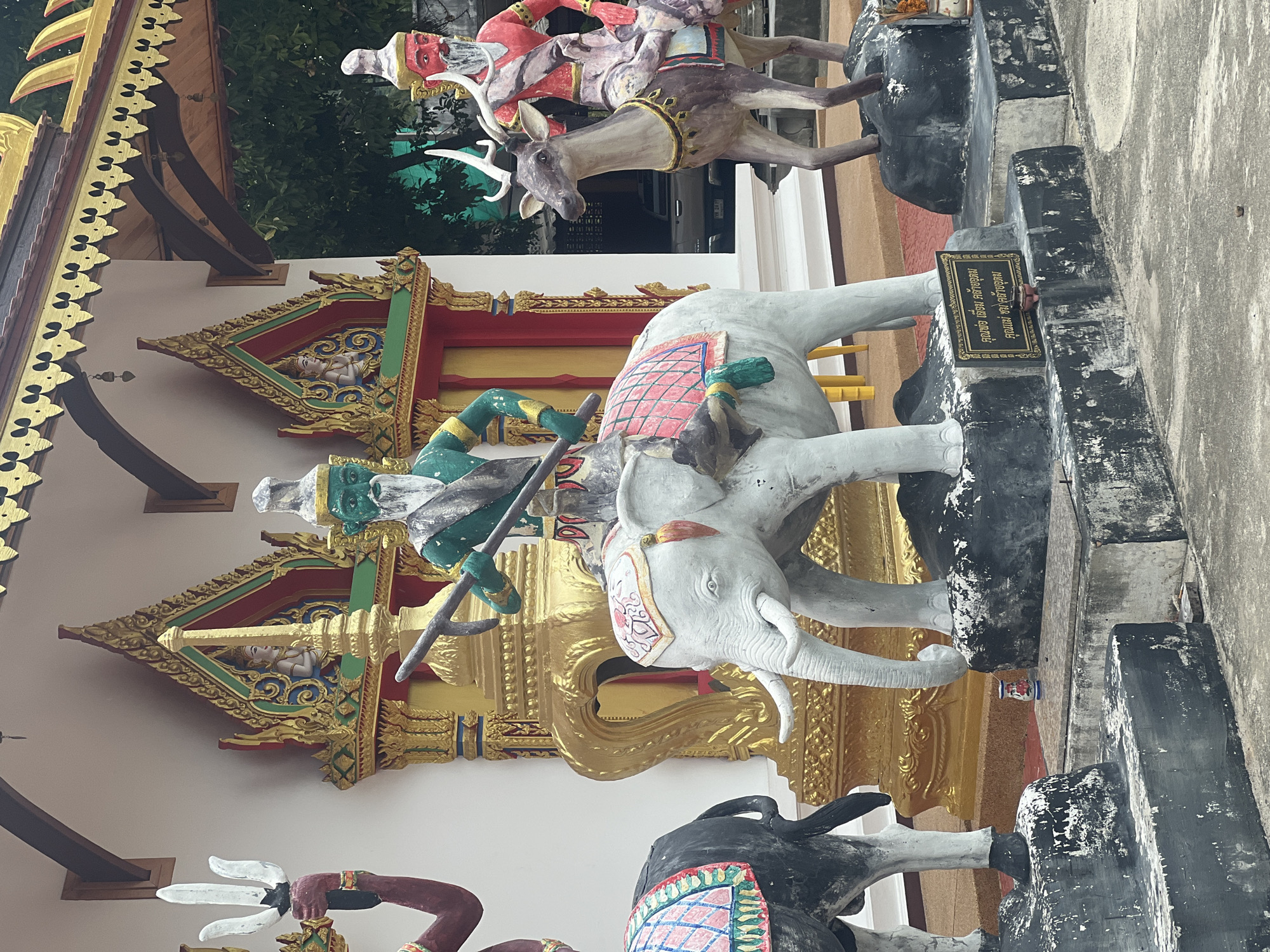 The image shows a religious statue display in front of a building with ornate golden trim and a dark roof. In the foreground, there are several statues. The most prominent statue is a white elephant with a person wearing green and a white beard, holding a weapon, mounted on its back. To the left of the elephant is a dark statue of an elephant decorated with a colorful saddle cloth. To the right of the white elephant is a statue of a deer with a person wearing red and a white beard, also mounted. All the statues are on dark, rocky bases. Behind the statues, the building has a white facade with golden decorative elements and red doors. There are also smaller figures in niches on the building. A sign with Thai text is visible in front of the white elephant statue. The ground in front of the statues is paved concrete.