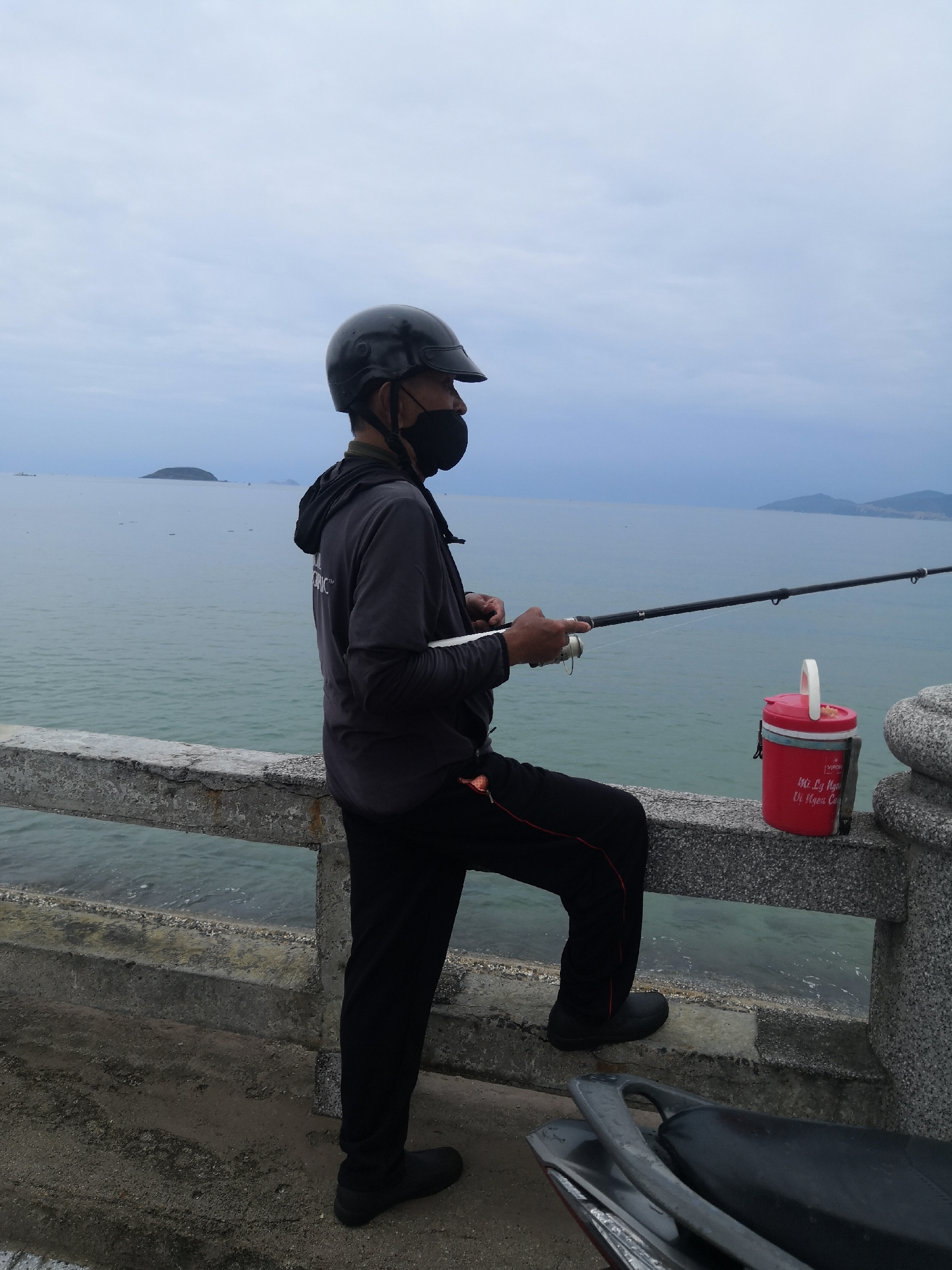 A person wearing a black helmet and face mask is fishing from a concrete pier in the city of Bac Nha Trang Ward, Vietnam. The person, dressed in a grey shirt and black pants, is holding a fishing rod and facing towards the sea. A red bucket is resting on the pier near the person. The water is calm with a few small islands visible in the distance. A part of a vehicle, possibly a motorcycle, can be seen in the foreground. The sky is overcast.