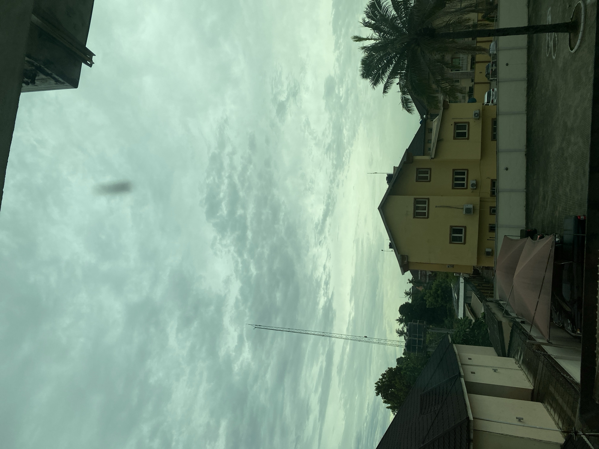 The image captures a partial cityscape in Ikeja, Nigeria, under an overcast sky with layered clouds. A tall, thin communication tower stretches upwards into the sky. A yellow building with multiple windows and air conditioning units is visible to the right, partially obscured by a palm tree. Below that, there's a building with a dark roof and a tan-colored exterior, and a pink outdoor umbrella. In the foreground, there are parts of buildings, and a patch of what looks like a parking lot. The image appears to be taken from an elevated perspective, likely from inside a building. A dark blob is visible in the sky, possibly a lens artifact.