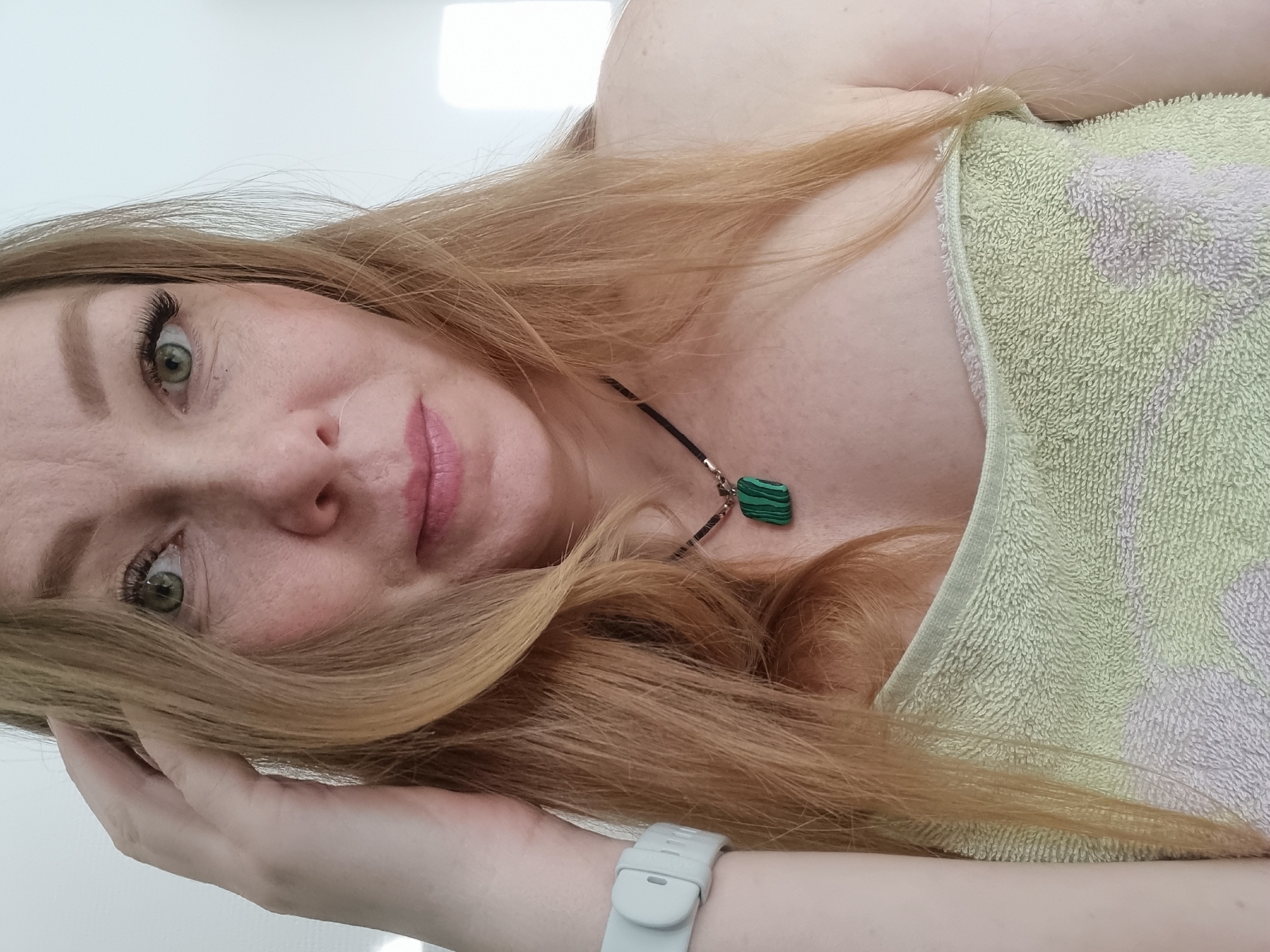 A close-up, angled portrait of a fair-skinned woman with long, light brown hair and green eyes, likely post-shower or bath. She is partially covered by a light green and white striped towel. She wears a necklace with a green pendant. A white fitness tracker is visible on her left wrist. Her left hand is positioned near her face, touching her hair. The background appears to be a plain white surface. No indication of the location is visible.