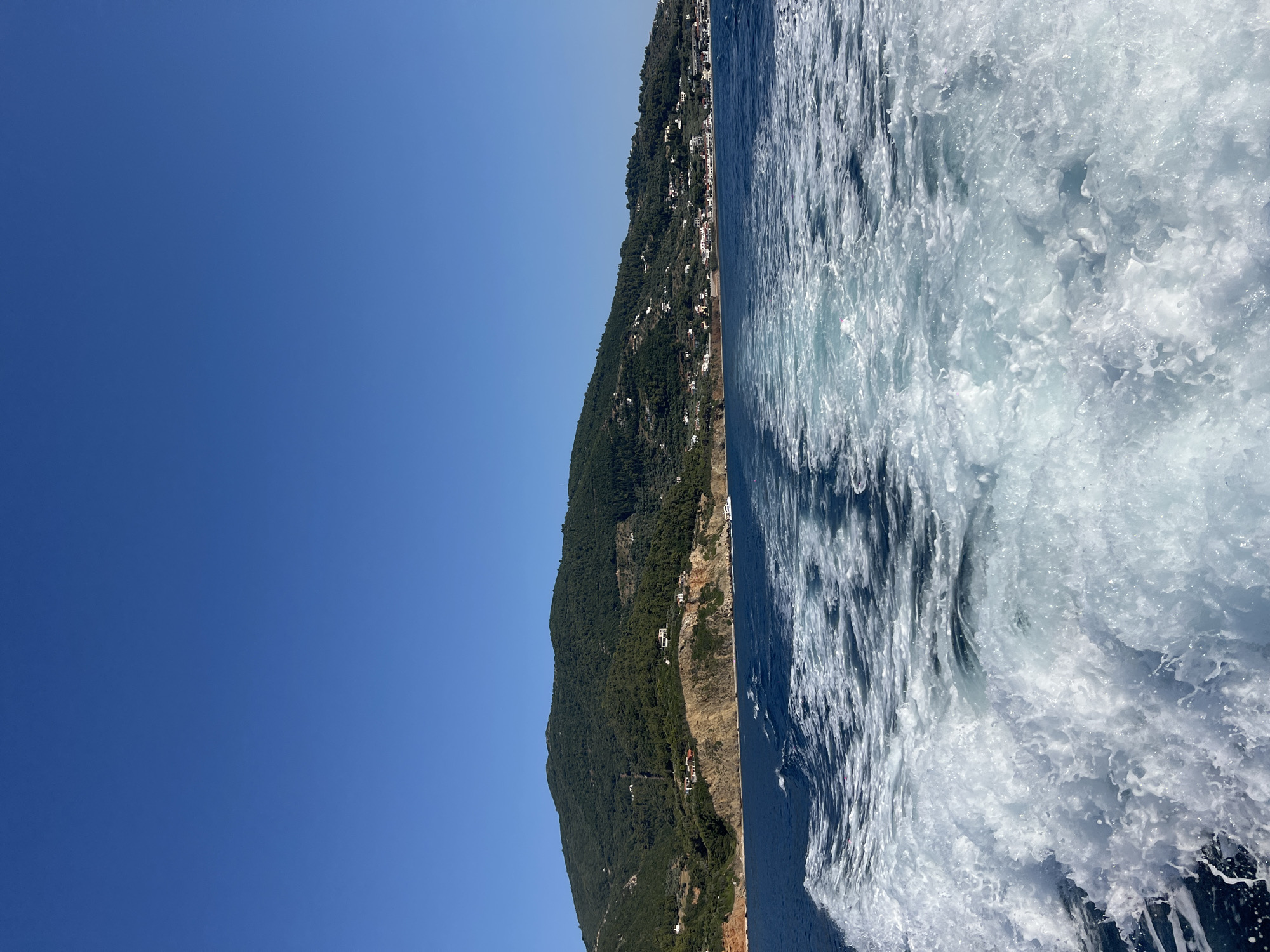 The image depicts a view from a boat moving through the Aegean Sea off the coast of Skopelos, Greece. In the foreground, white foamy wake churns the deep blue water. The middle ground shows the clear blue sea stretching towards a verdant, hilly coastline. Along the slopes of the hills, small clusters of white buildings, characteristic of Greek island architecture, are visible. The coastline is a mix of rocky outcrops and vegetated slopes, with trees and olive groves dotting the landscape. The background is dominated by a clear, bright blue sky. The scene conveys a sense of movement and a coastal vacation atmosphere.
